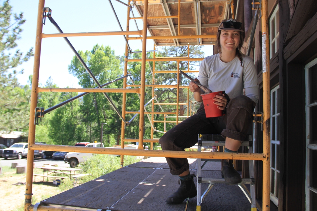 A person sitting on a scaffold holding a paintbrush and red bucket, with trees and parked cars in the background. Scaffolding surrounds the person, who is wearing a hat and work clothes.