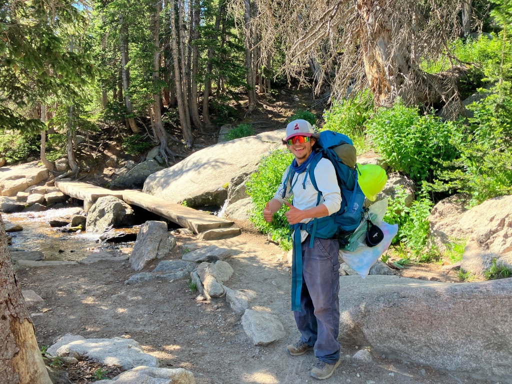 A person in hiking gear stands on a rocky forest trail. They are carrying a large backpack and smiling. A small stream and a wooden footbridge are visible in the background.