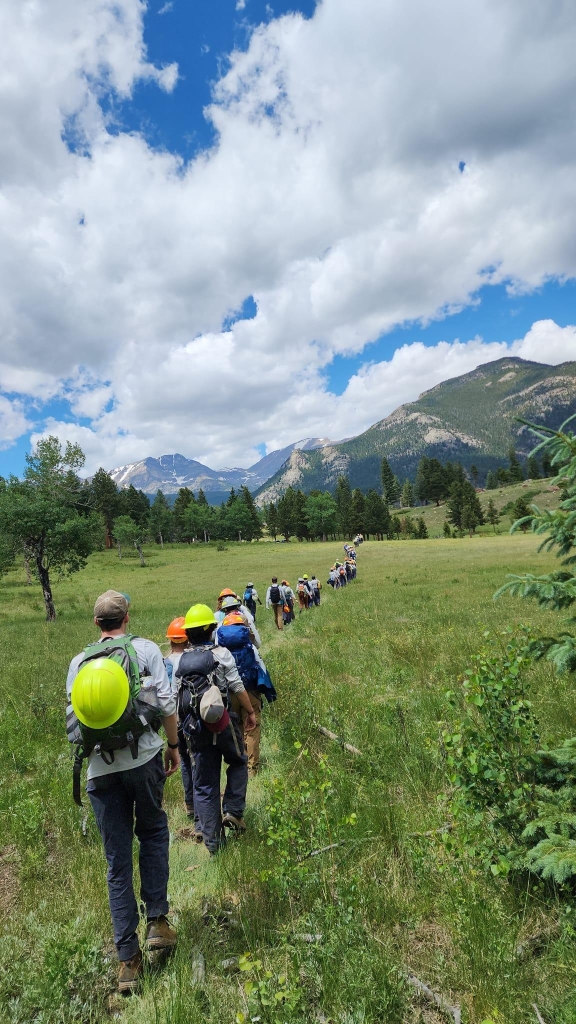 A group of hikers with backpacks and helmets walks in a line through a grassy meadow with mountains and a partly cloudy sky in the background.