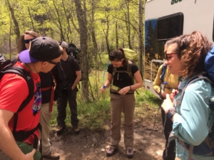 A group of hikers with backpacks listens to a guide demonstrating something on the ground