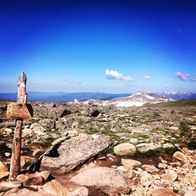 Wooden trail sign on a rocky mountain path with expansive blue sky and distant snow-capped peaks.