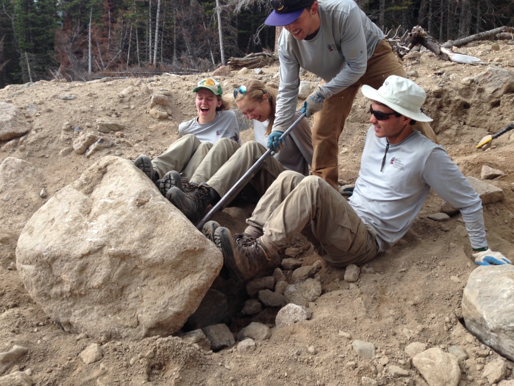 Four people in workwear laugh while pushing a large rock