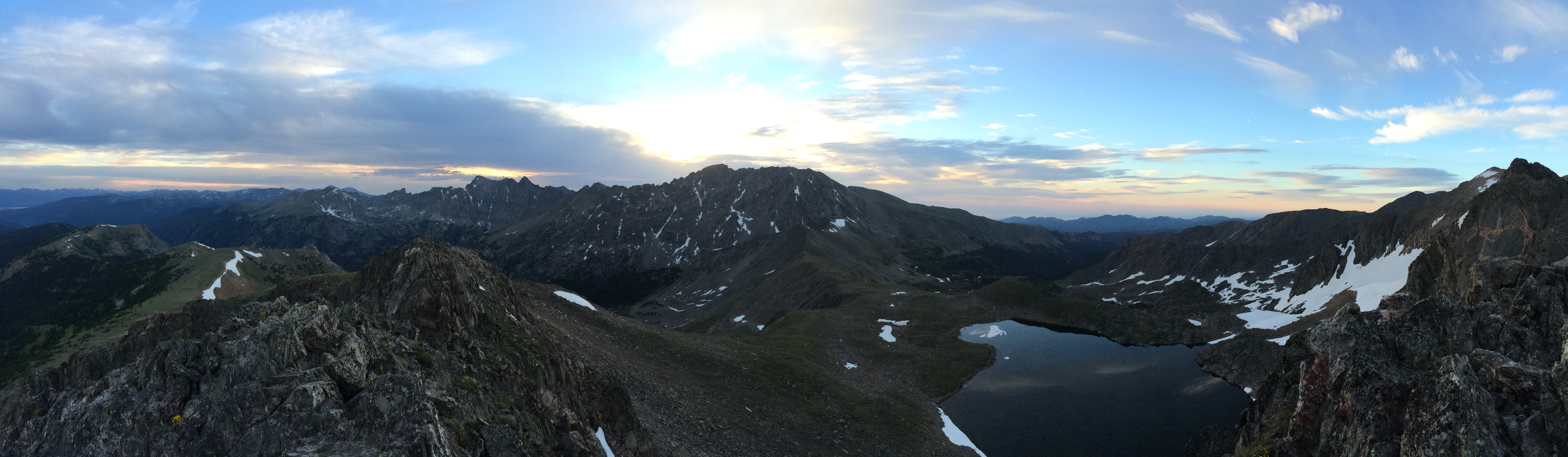 Panoramic view of rugged mountains with small snow patches and lakes at sunset, under a vast sky.