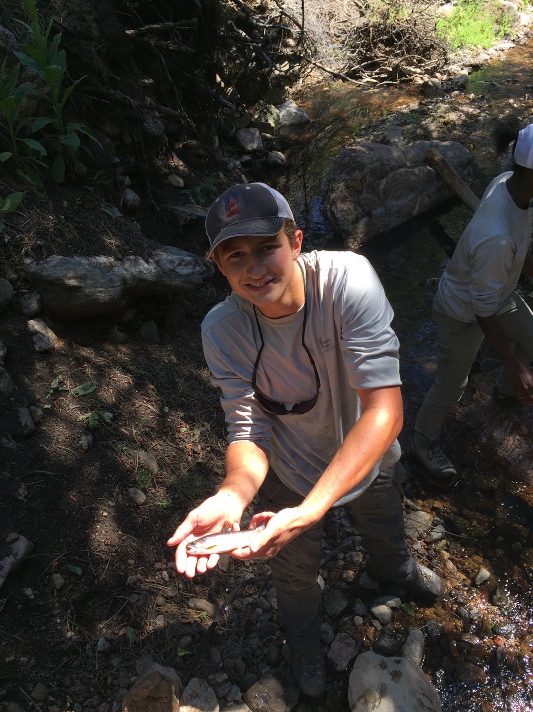 A man in a cap and long-sleeve shirt smiling at the camera, holding a small fish