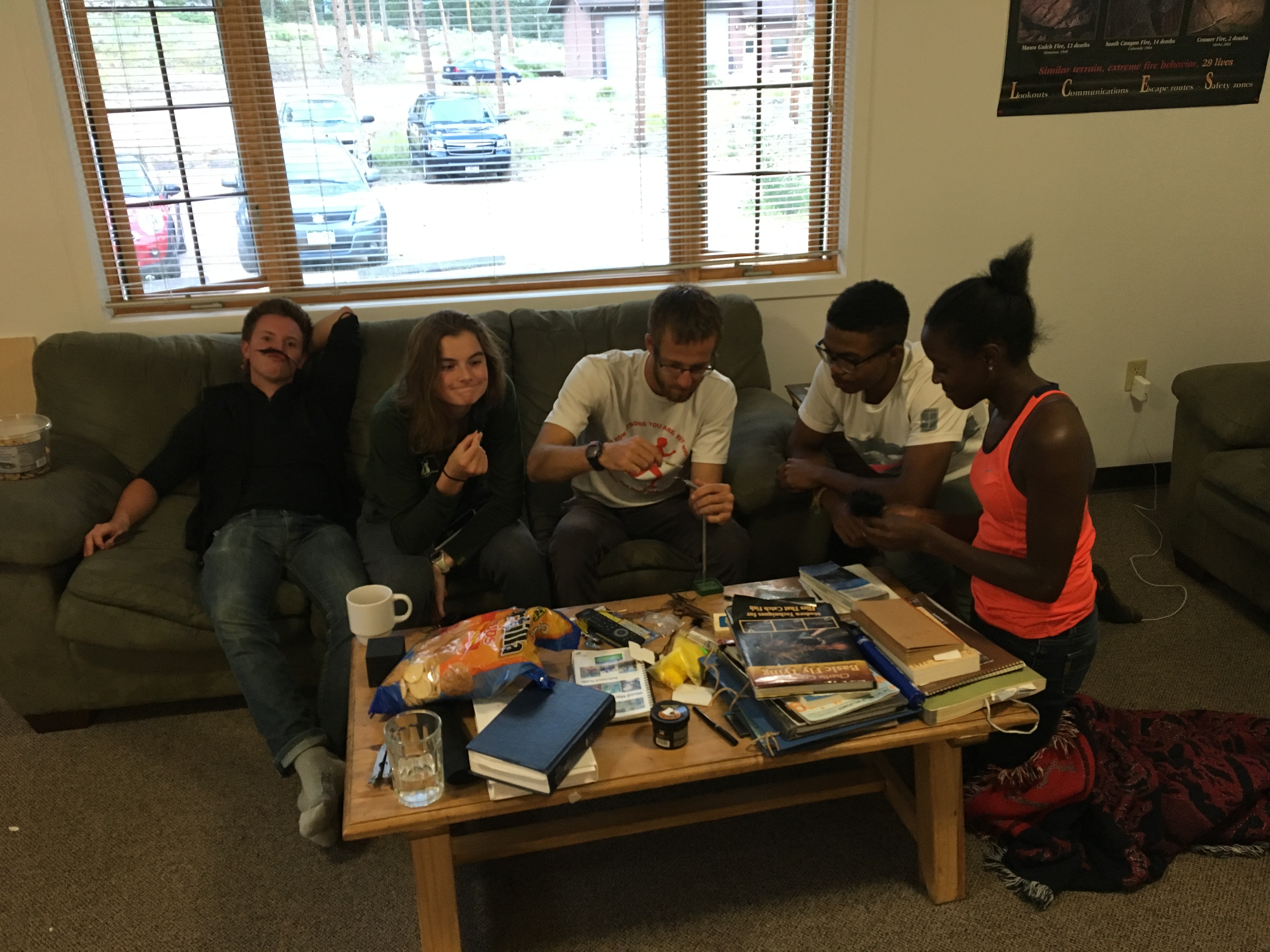 Five people sitting on a couch, interacting and surrounded by snacks and magazines