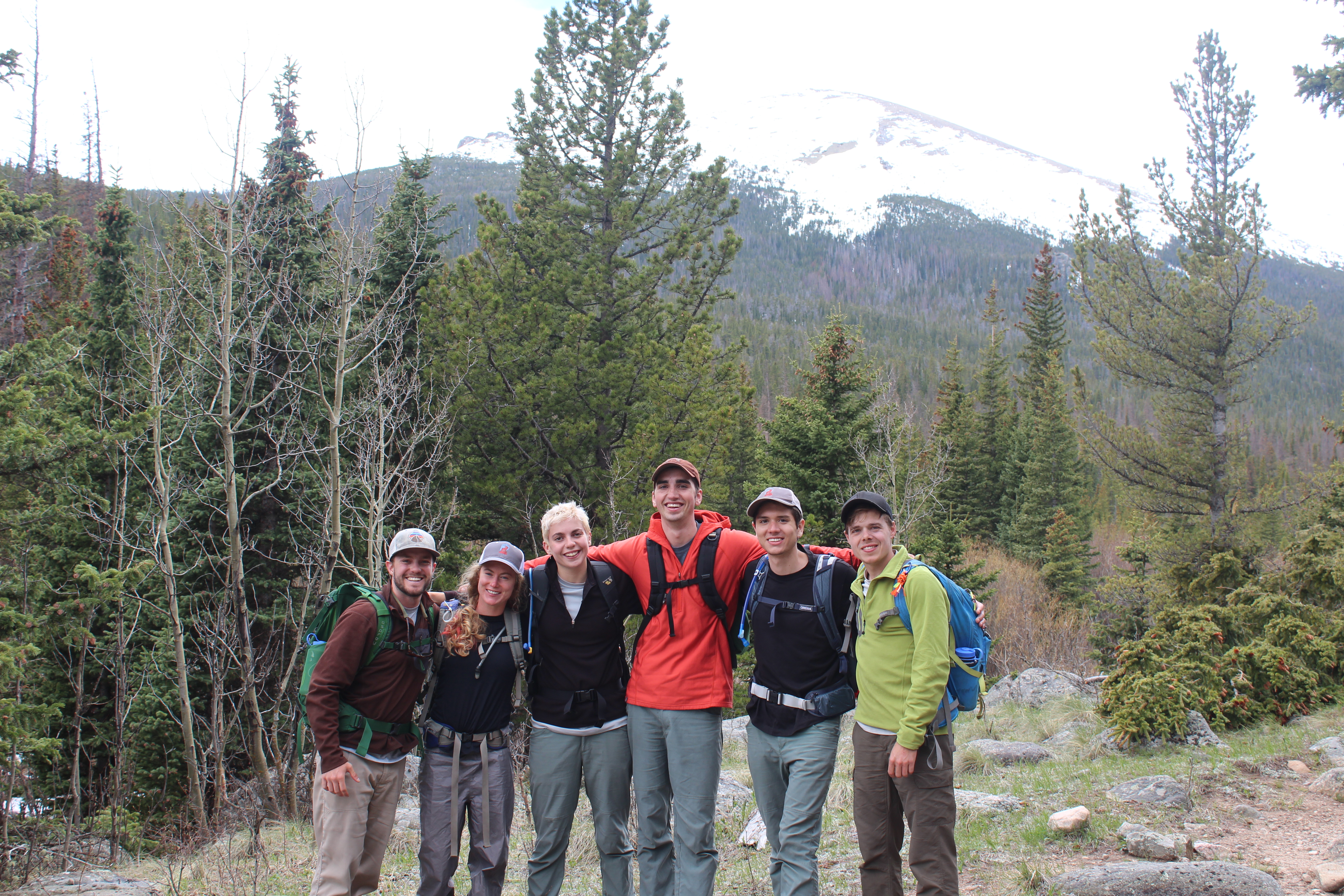 Group of six hikers smiling in front of a forest with a snow-capped mountain in the background.