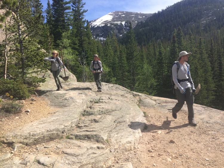 Three hikers with backpacks walk on a rocky trail in a forested area