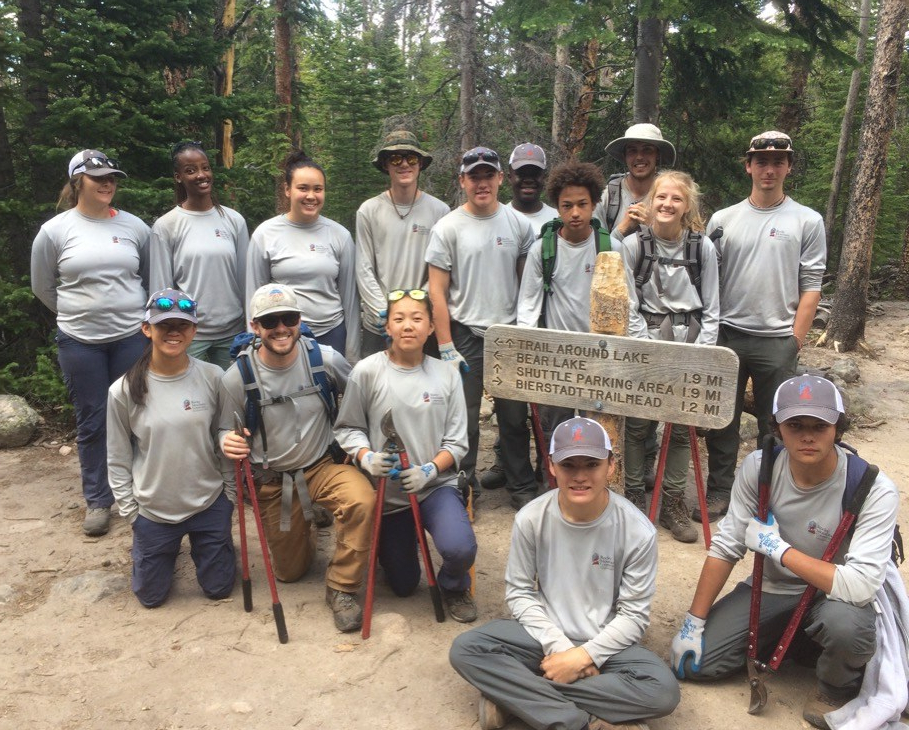 Group of diverse hikers posing with trekking poles next to a trail sign in a forest.