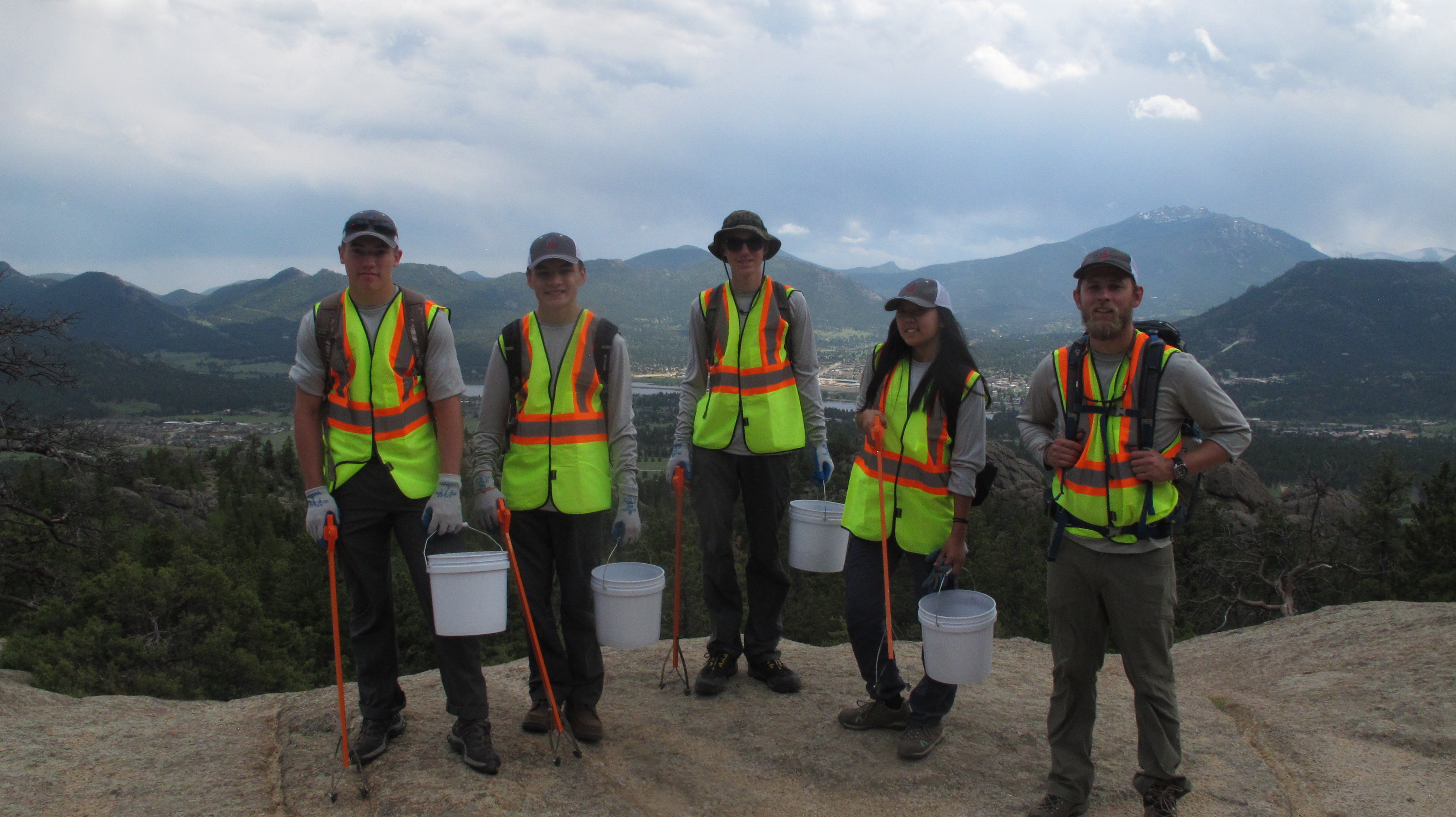 Five people wearing reflective vests and holding buckets stand on a rocky overlook