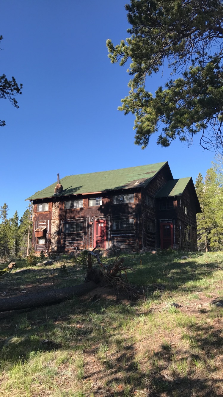 A large rustic log cabin in a forest setting with sunny skies and scatted trees around.