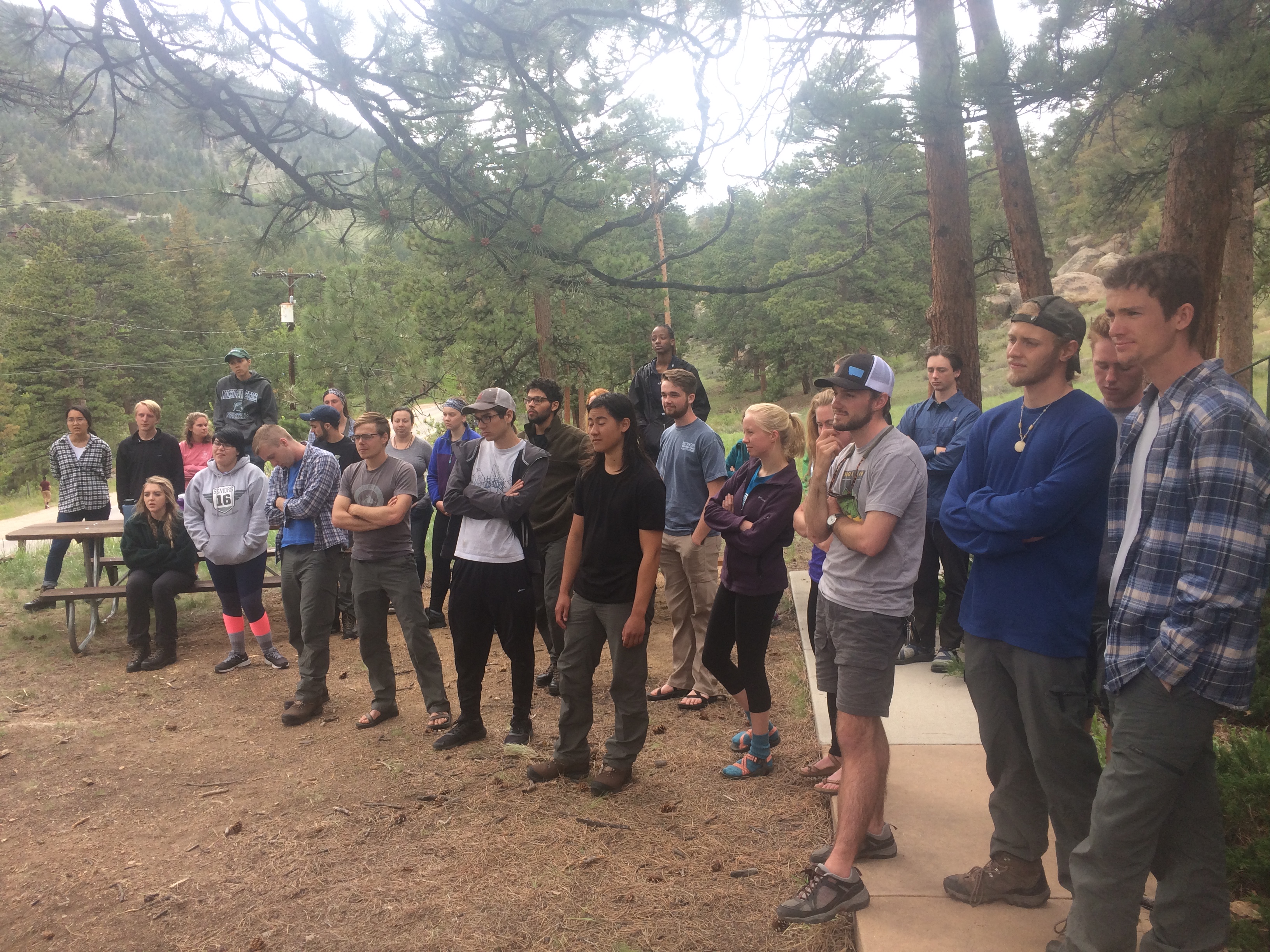 Group of people standing outdoors in a forested area, listening attentively to a speaker