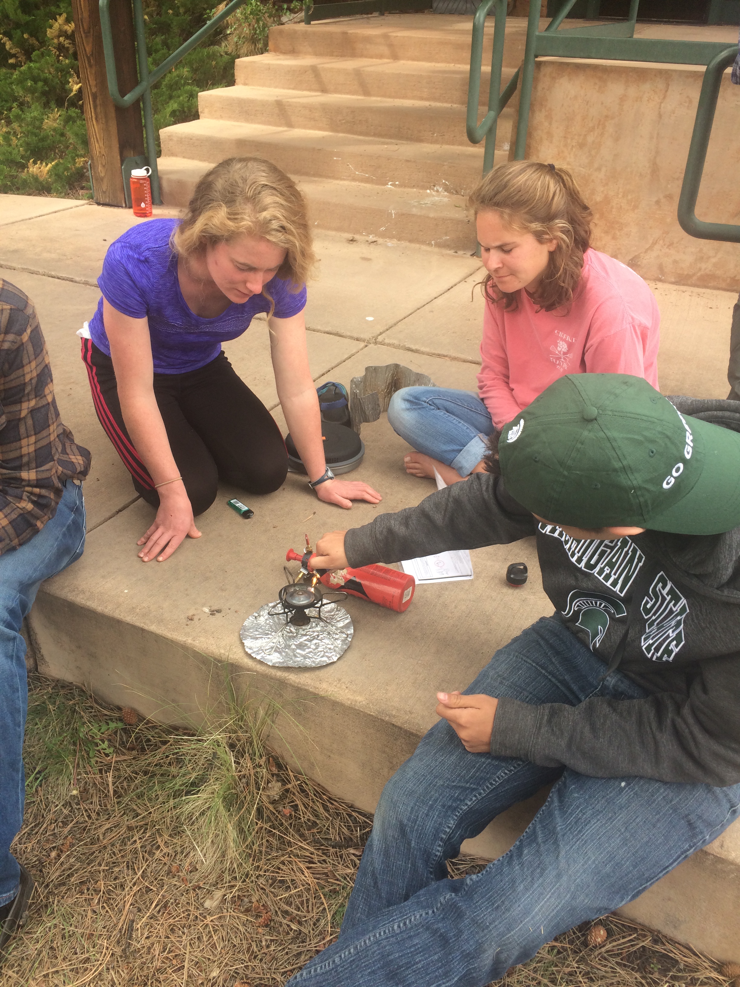 Three people using a portable stove on a concrete step outdoors