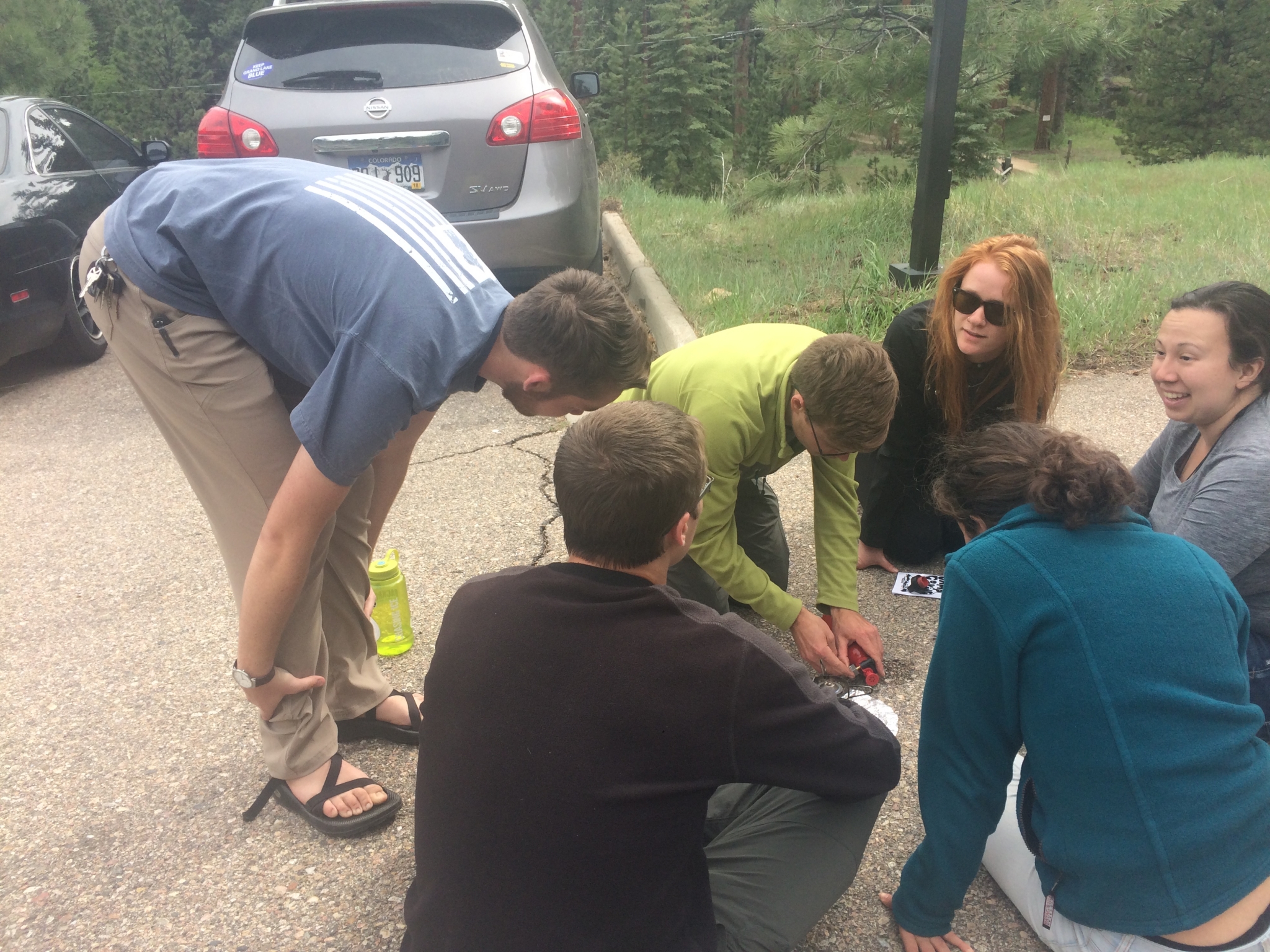 Group of people examining a tire pressure gauge together in a parking lot beside two parked cars.