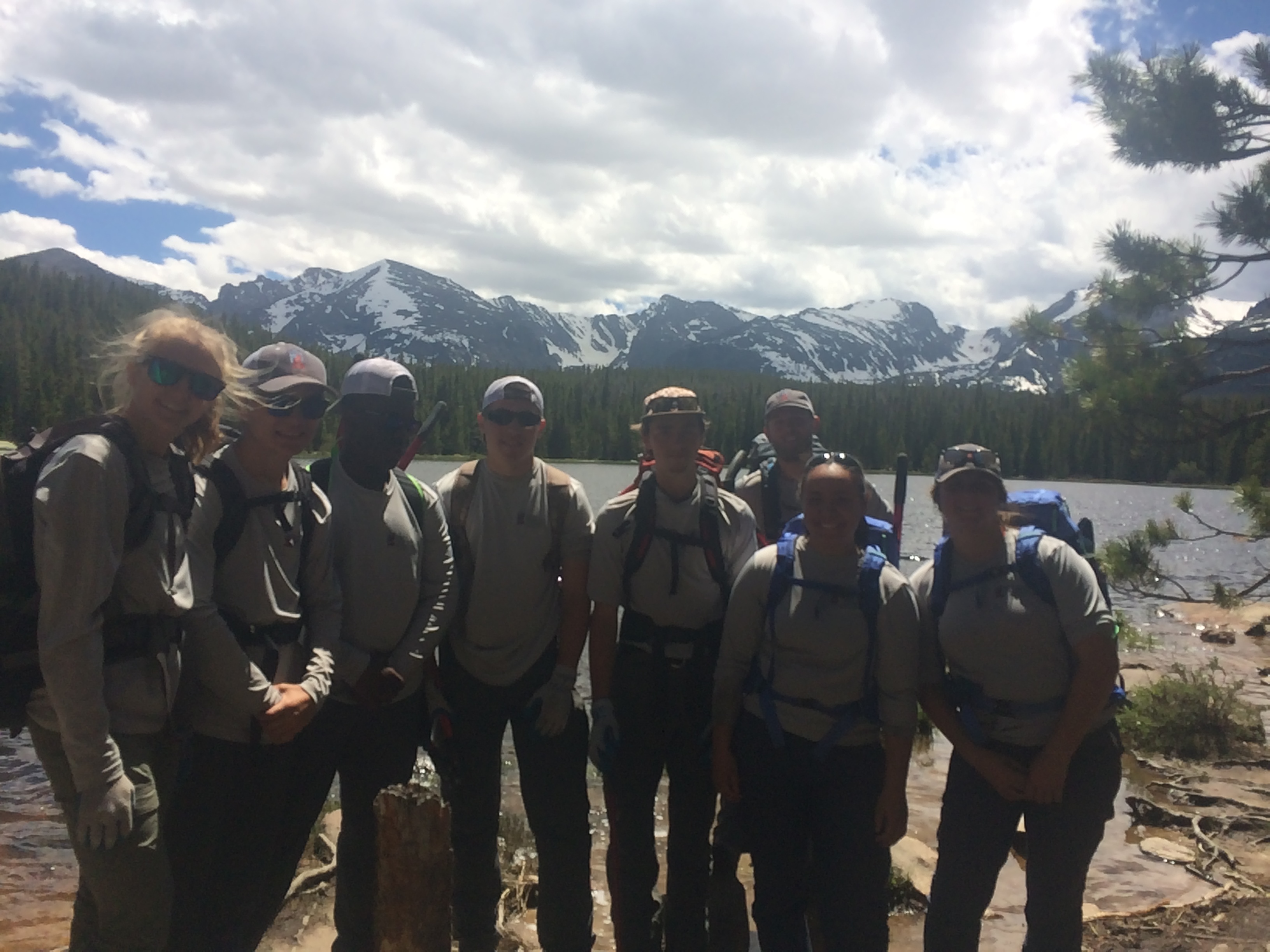 Group of hikers posing in front of a scenic mountain lake under a cloudy sky.