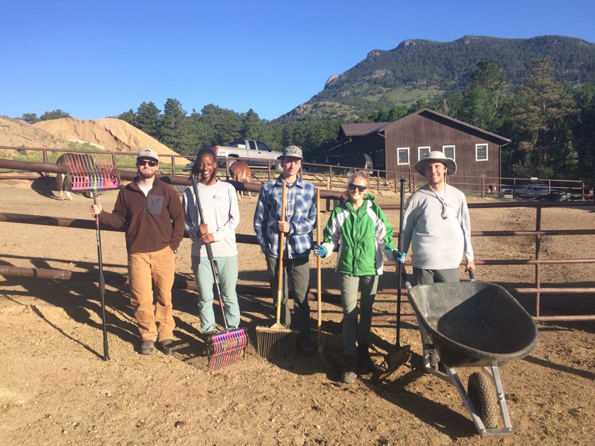 Five people posing with farm tools in front of a corral and a mountain.