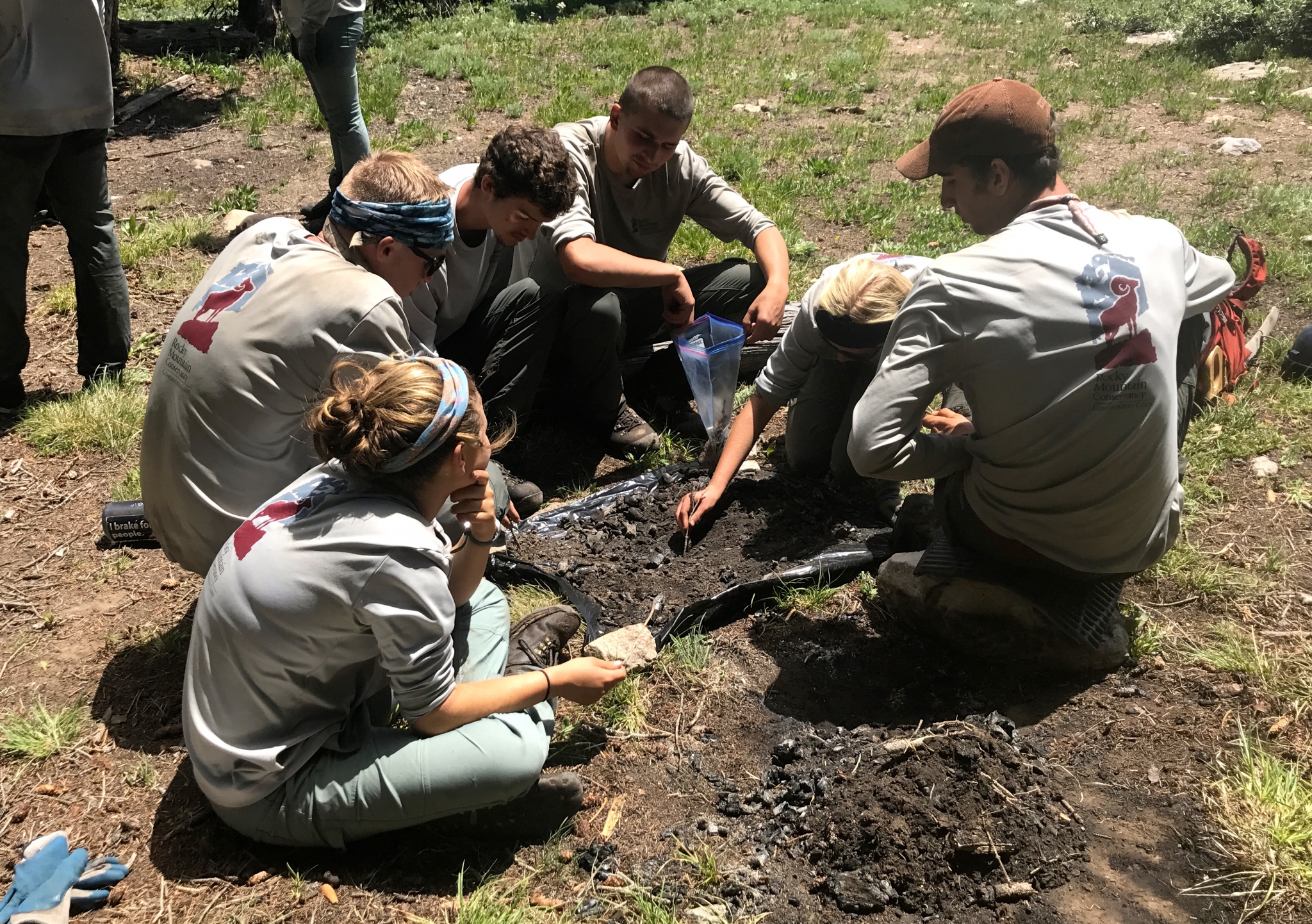 A group of young adults in outdoor attire gathers around a dirt site, engaging in a hands-on environmental study or activity in a forested area.