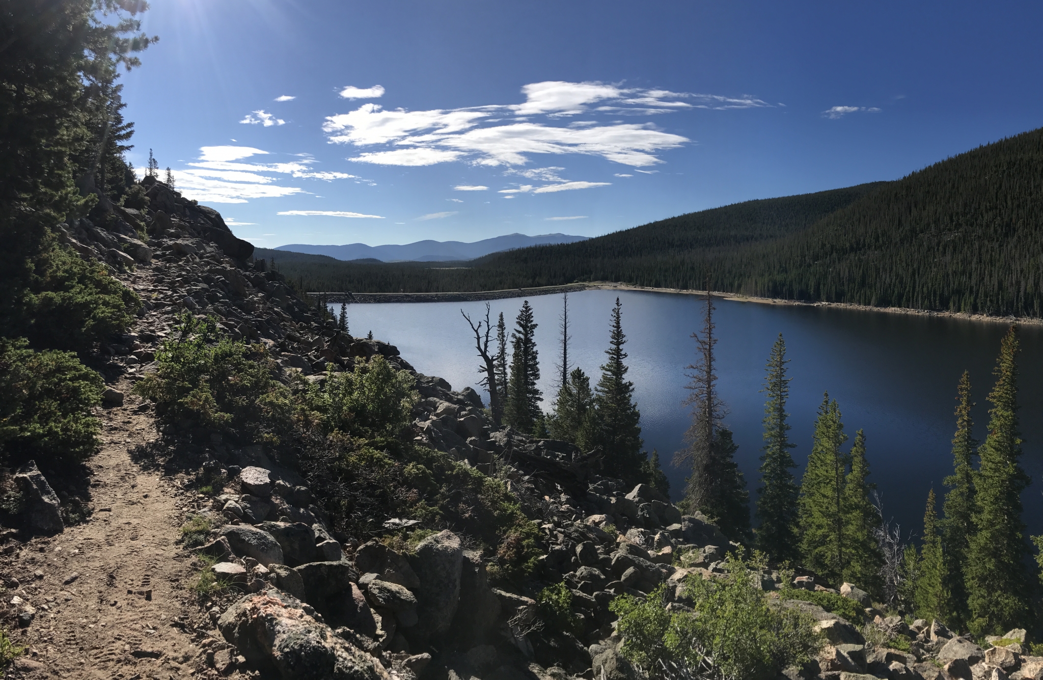 Panoramic view of a tranquil lake surrounded by lush pine forests under a clear sky with scattered clouds. a rocky trail runs along the lake's edge.