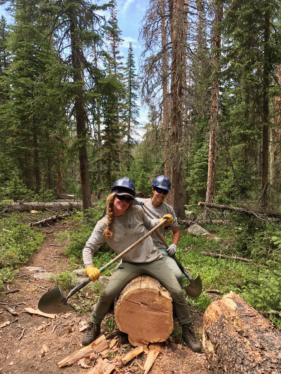 Two people in helmets sitting on a large log in a forest, holding shovels and smiling at the camera.