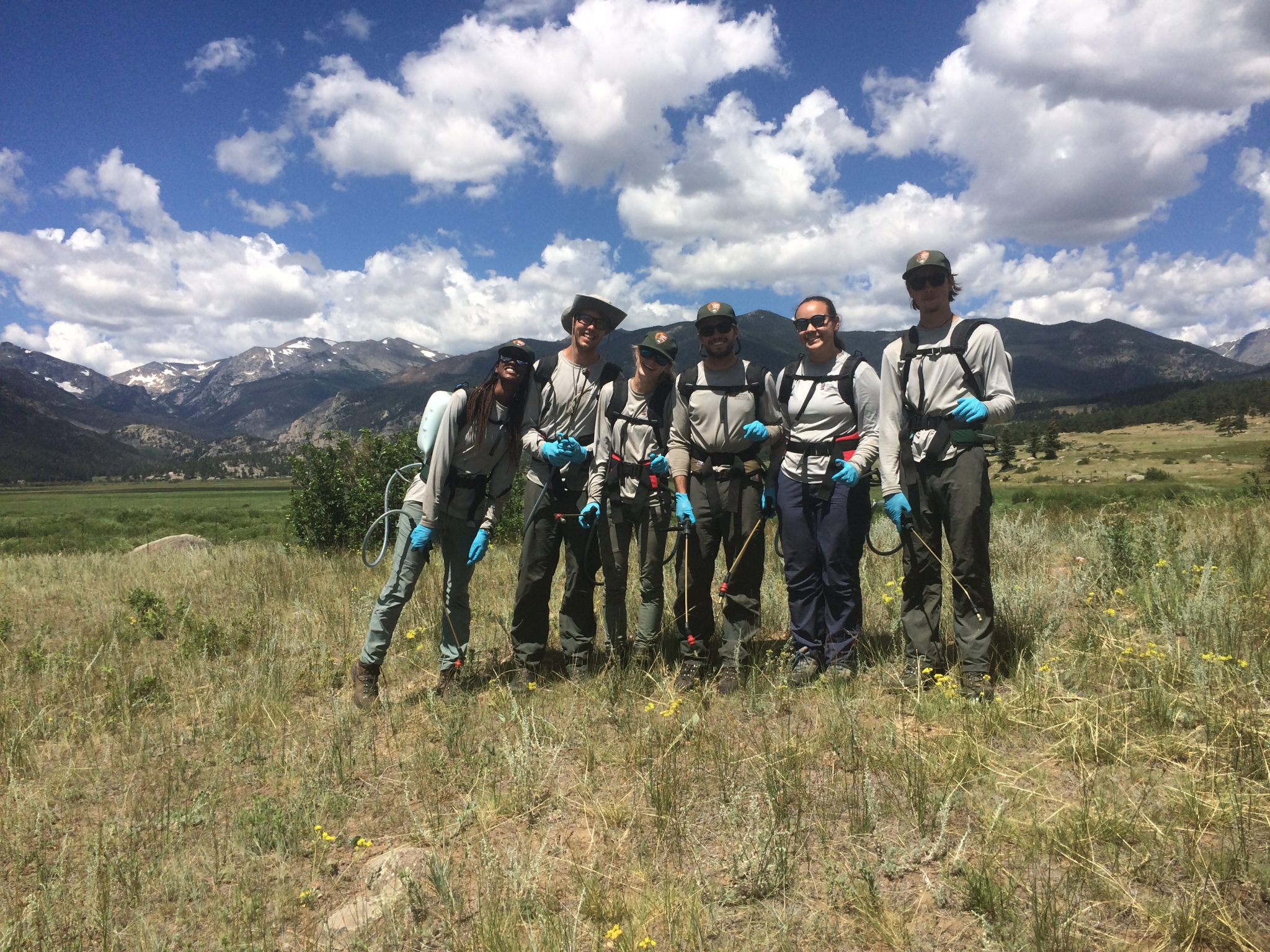 Group of six people in hiking gear with backpacks posing in a field with mountains in the background.