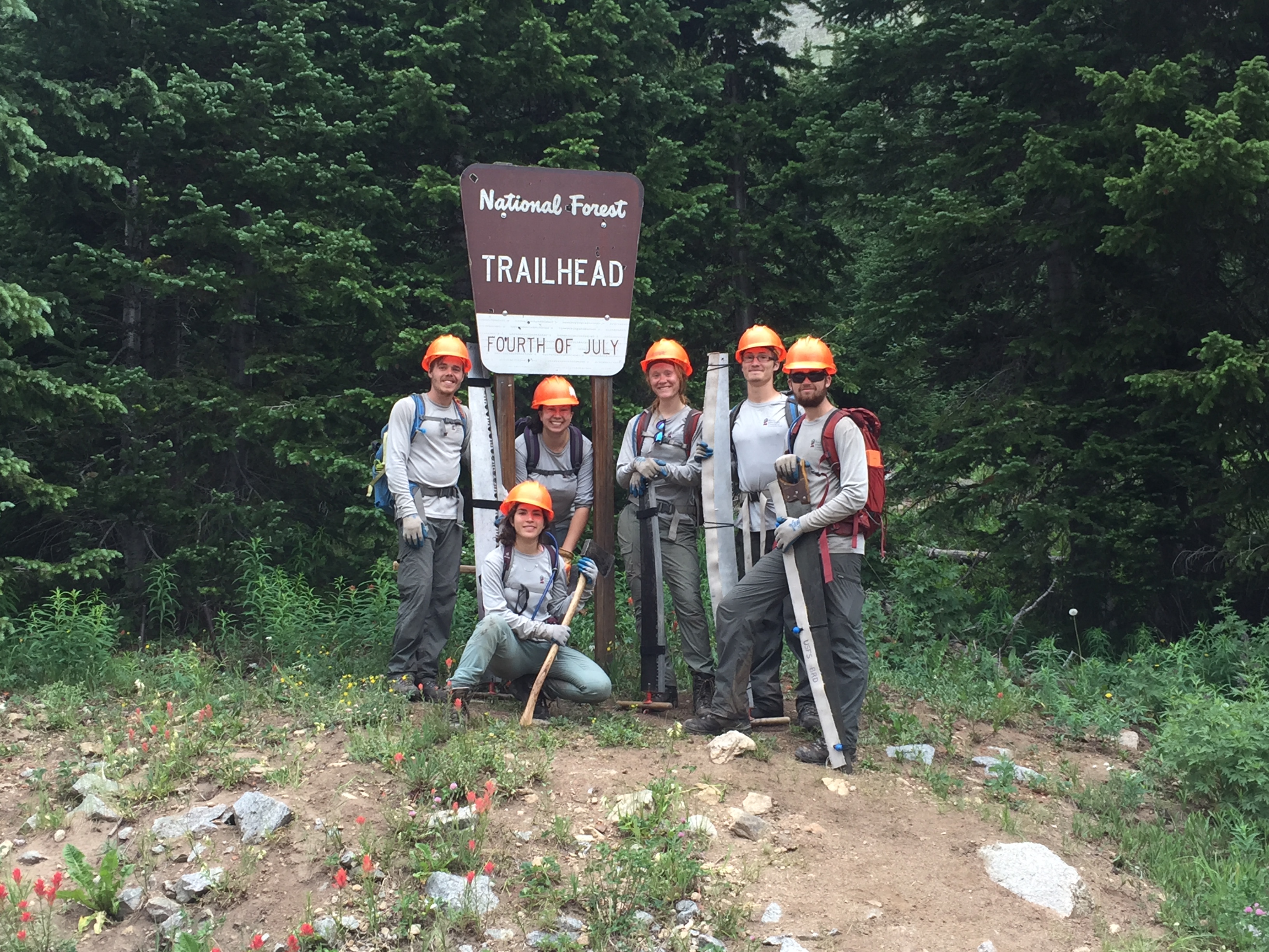 Six people wearing orange helmets pose at the "fourth of july trailhead" sign in a forested area.