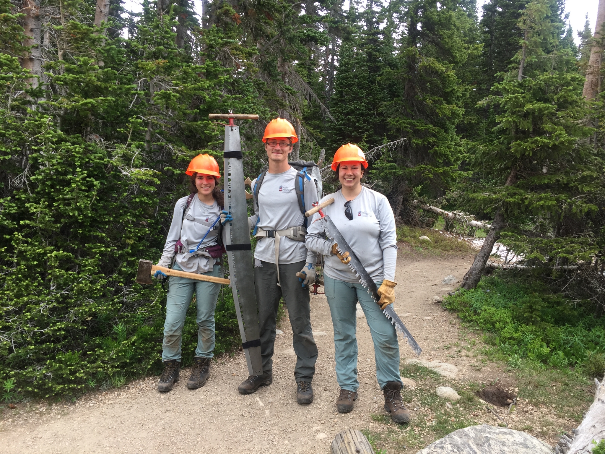 Three people in hard hats and work gear posing with trail maintenance tools in a forested area.