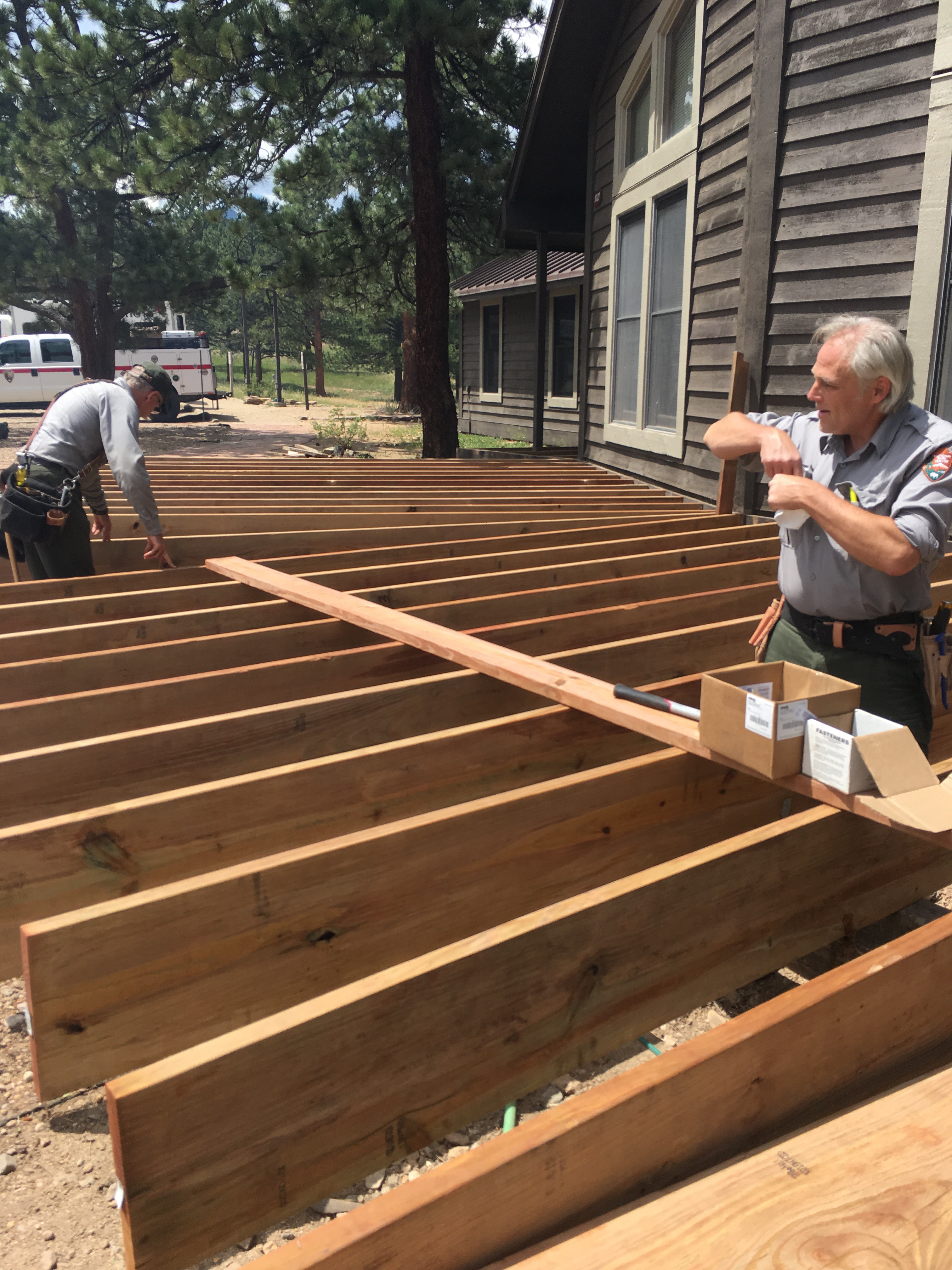 Two men constructing a wooden deck, measuring and securing beams next to a building.