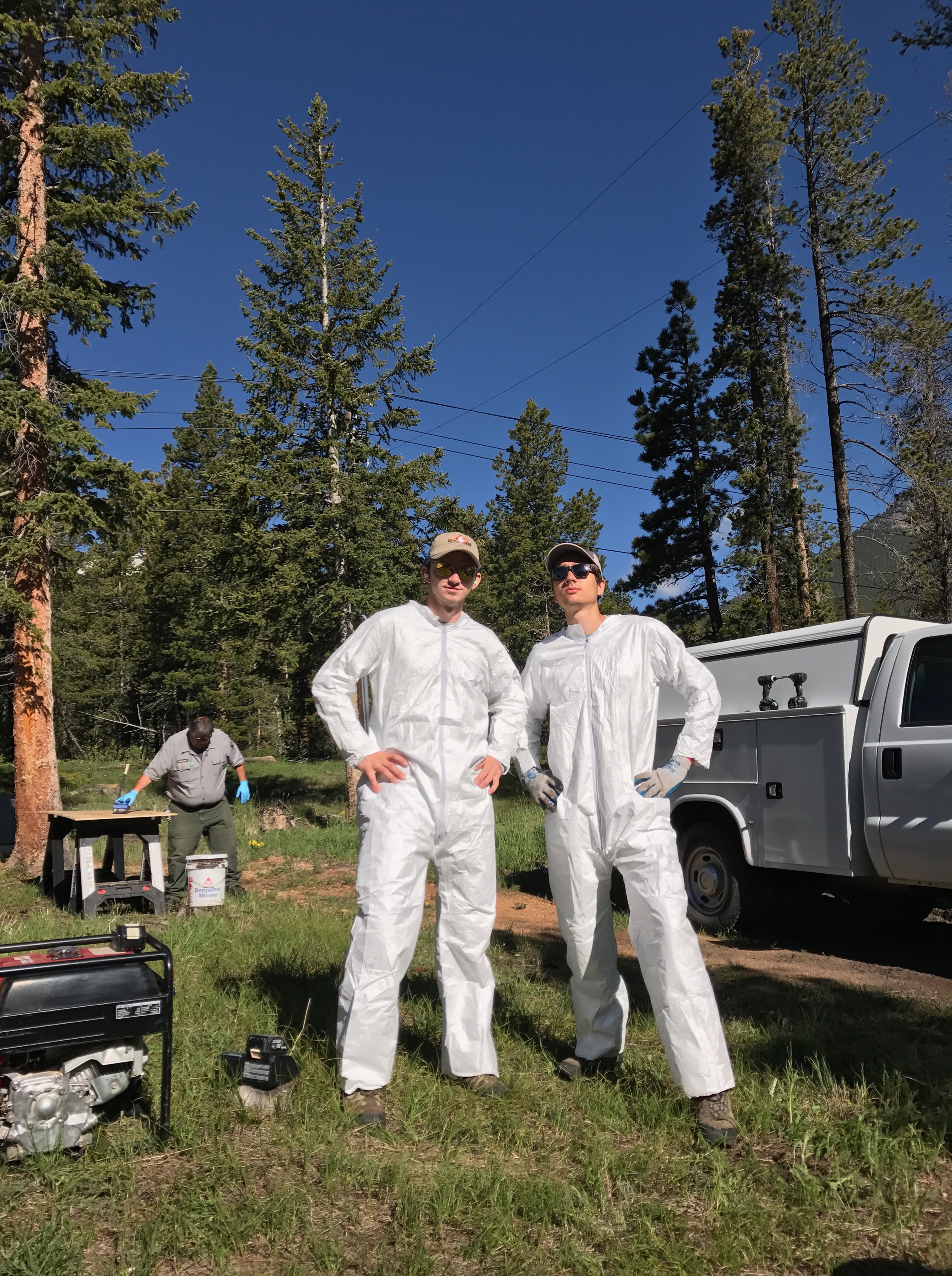 Two people in white protective suits and sunglasses stand confidently in a forest setting, with a truck and another person in the background.