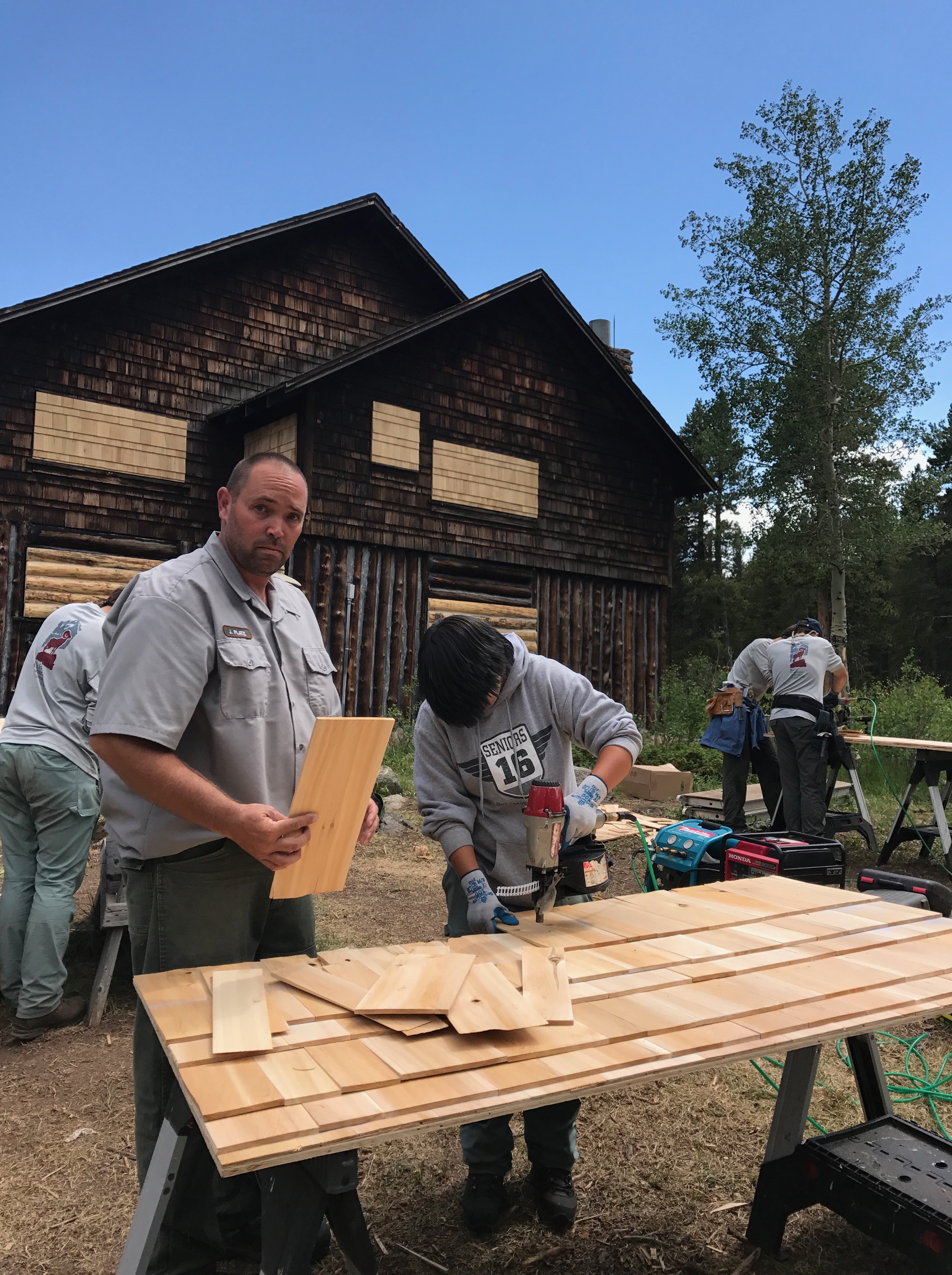 Man observing another man cutting wood on a table at a construction site with others working in the background near wooden cabins.