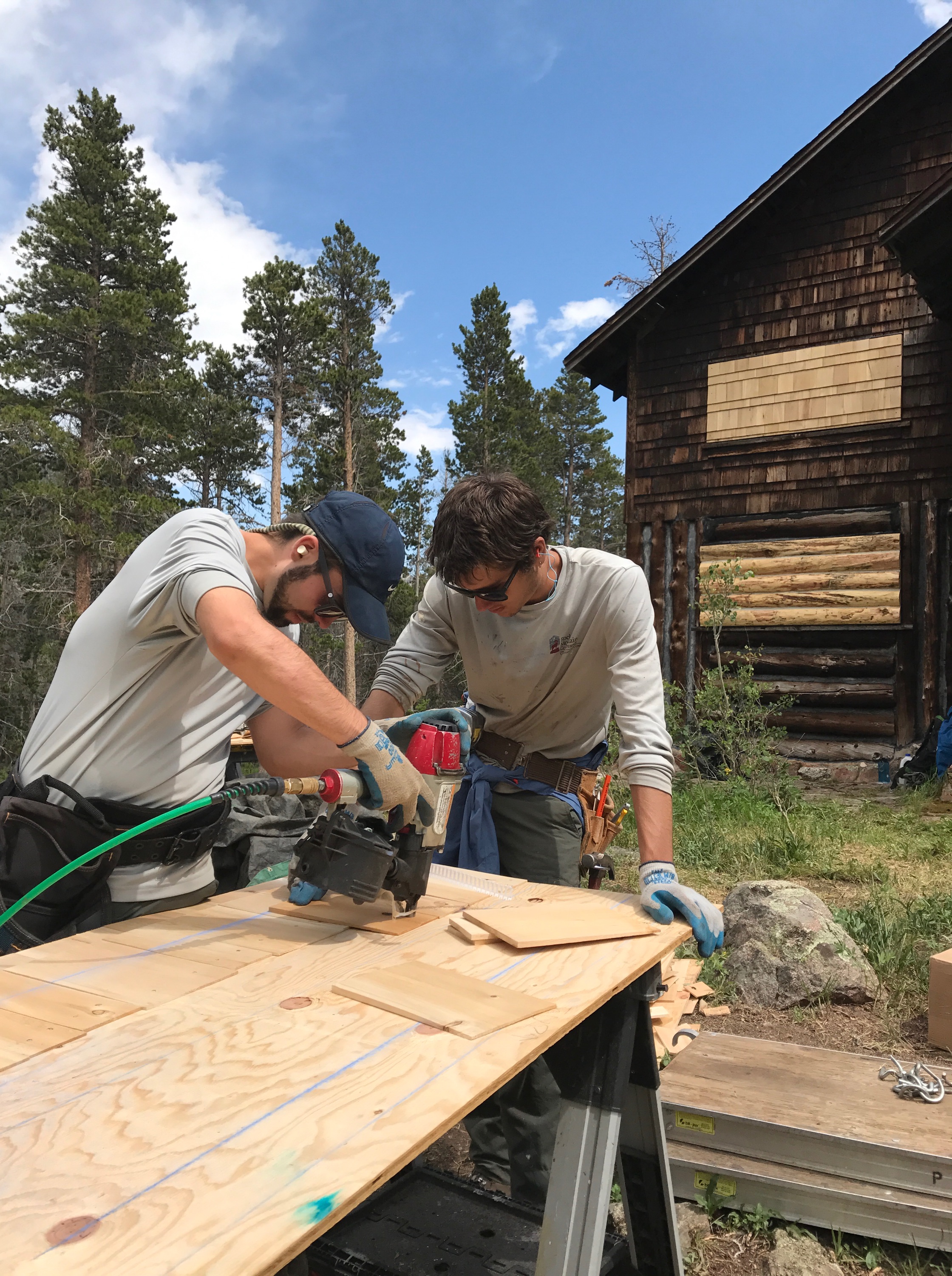 Two men using a circular saw to cut a wooden board on a worktable outdoors, with a rustic cabin and trees in the background.