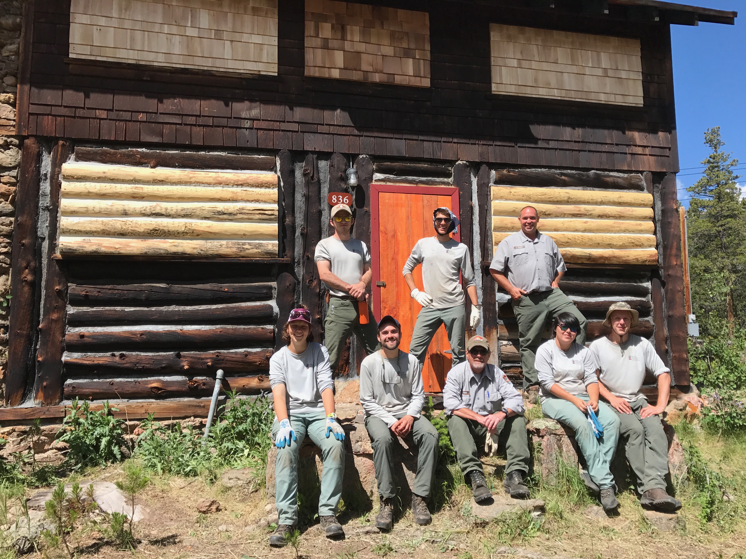 Eight park rangers in uniform posing in front of a log cabin with a red door, surrounded by greenery under a clear sky.