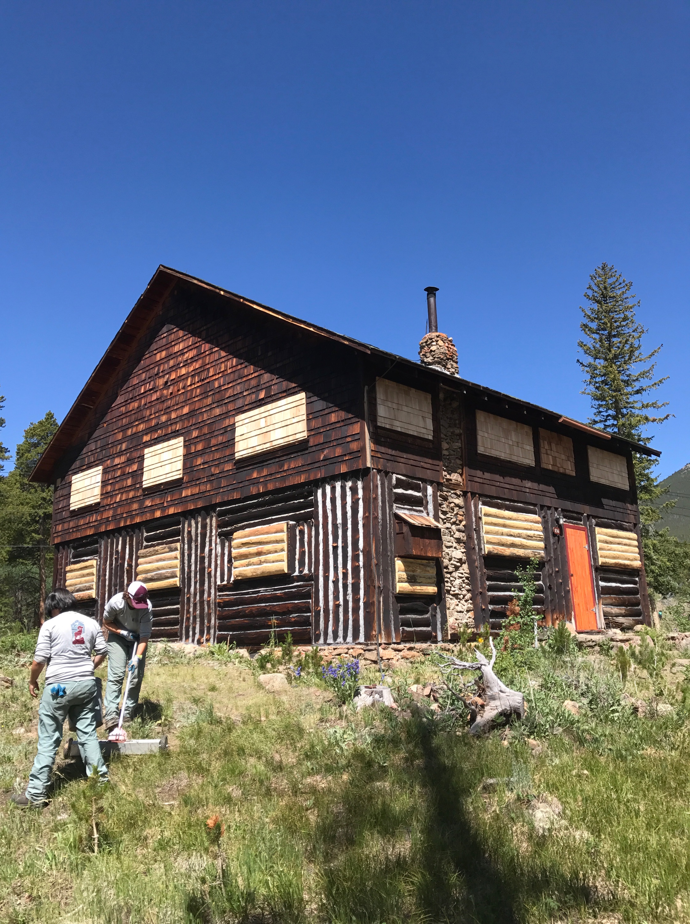 Two people gardening beside a rustic log cabin in a wooded mountain area under a clear blue sky.