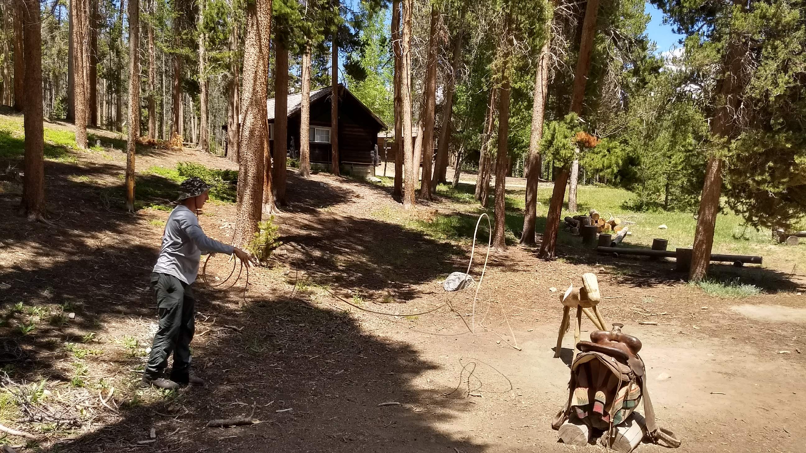 A person in a hoodie and hat uses a rope near a cabin in a sunny forest, with a backpack and walking stick in the foreground.