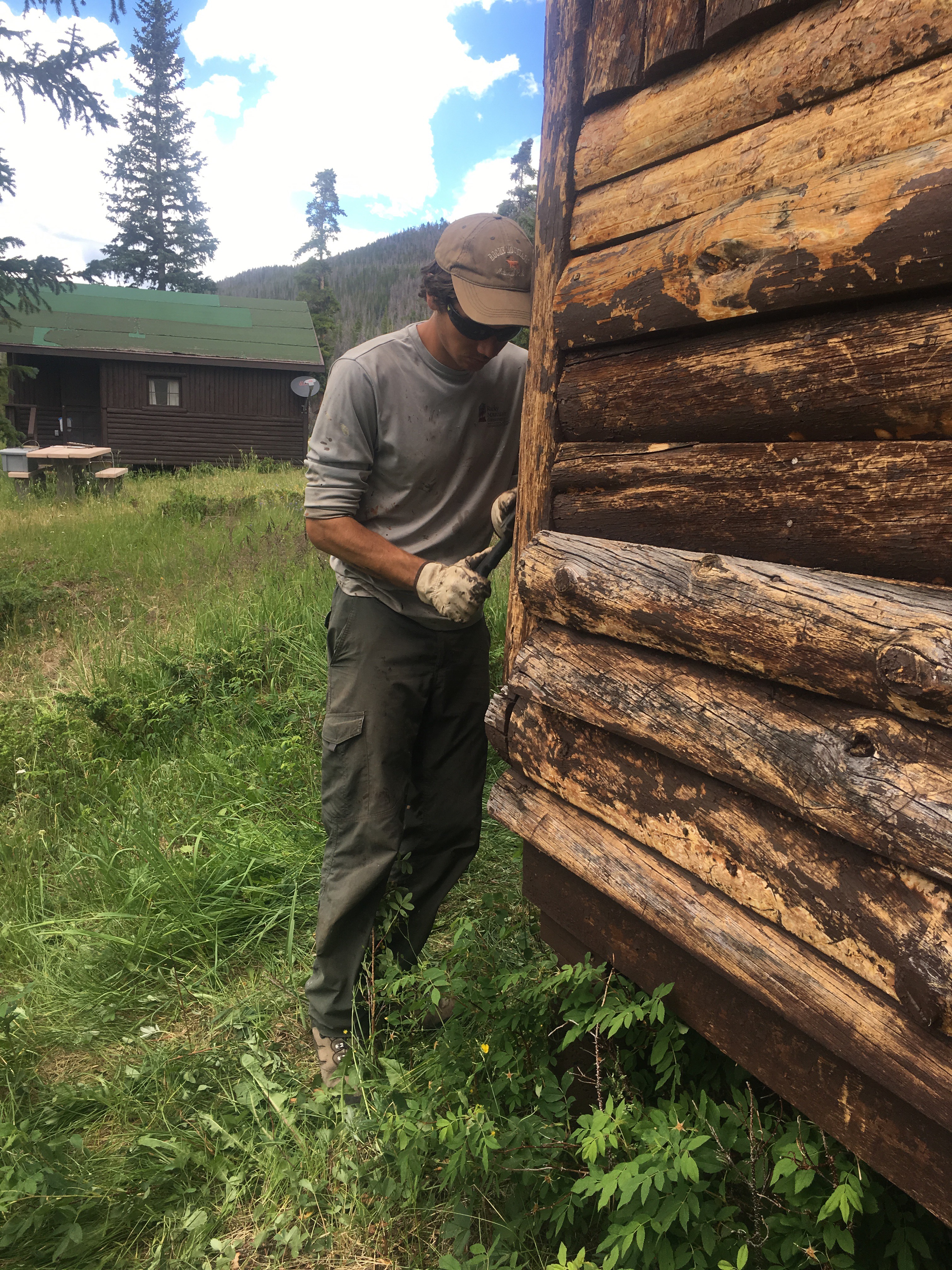 A man in work gloves scrapes the wall of an old log cabin