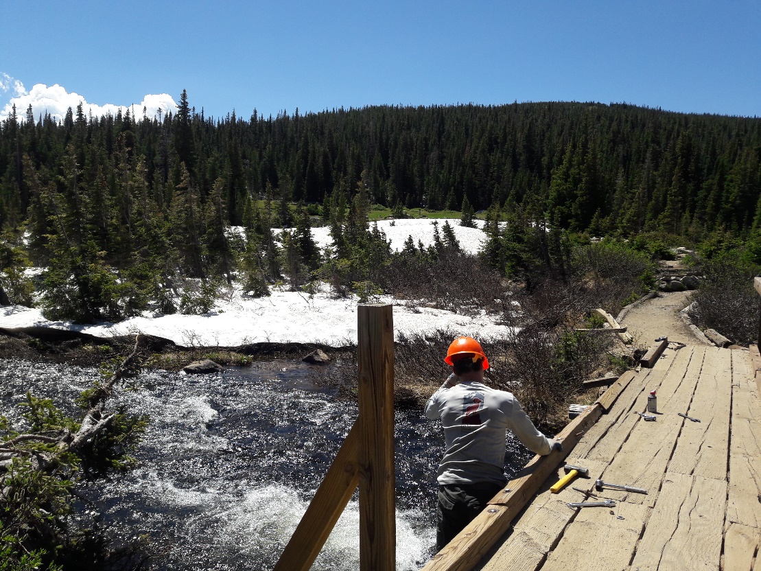 A person in a helmet crouches on a wooden bridge over a rushing river