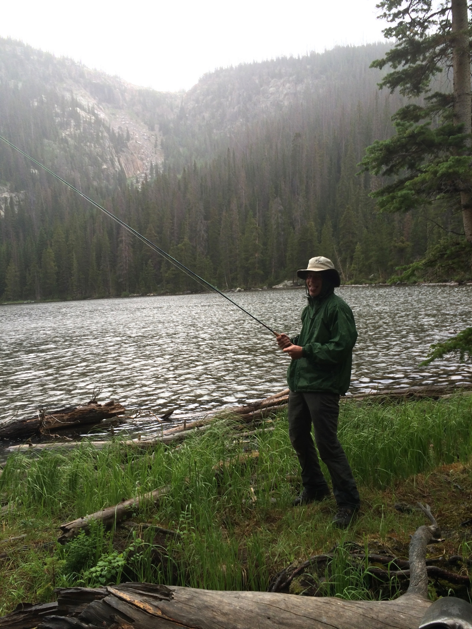 A man in a green jacket and hat fishing at a forest-lined lake under overcast skies.