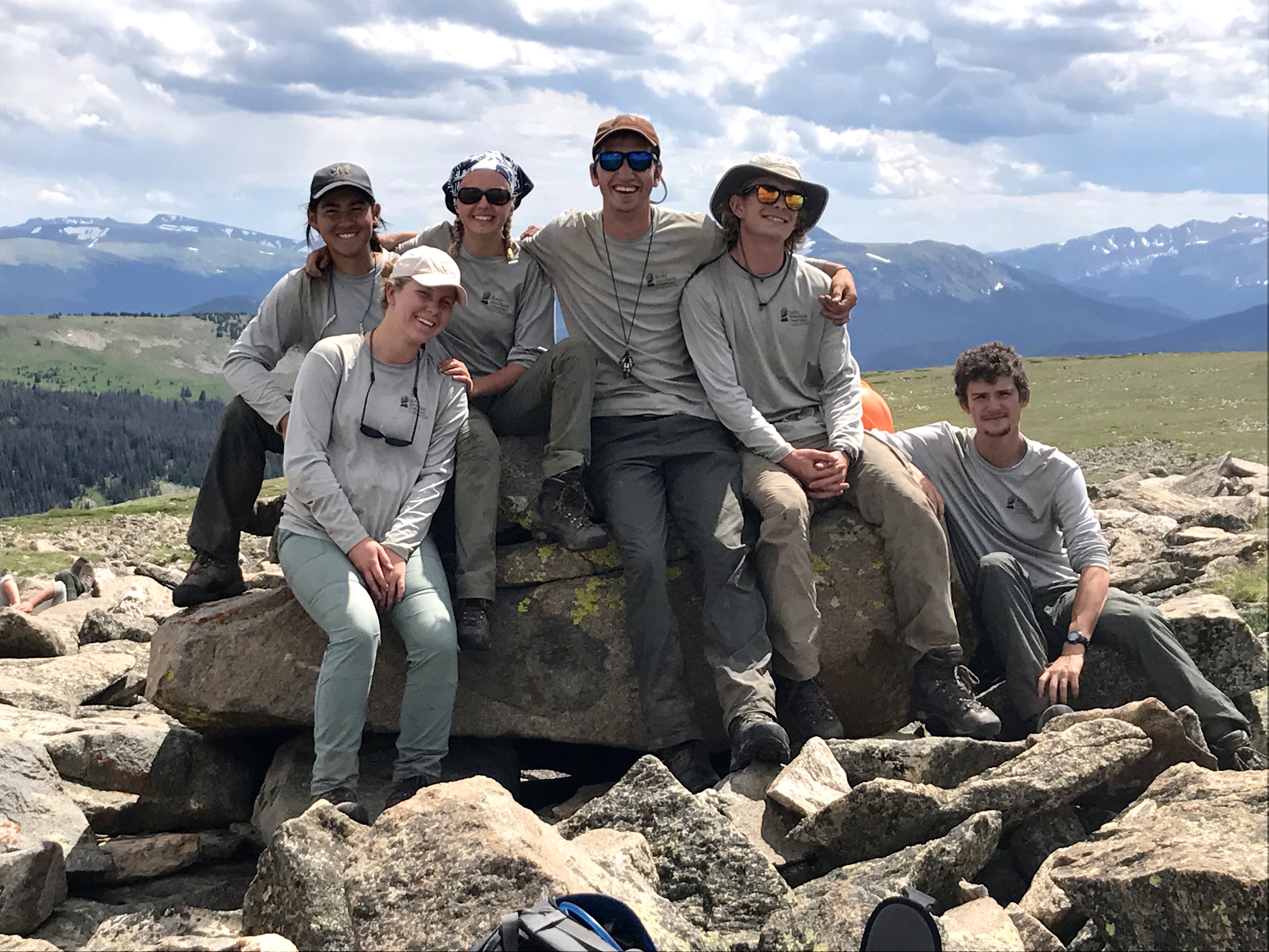 Six hikers posing on a large rock with a mountainous backdrop under a cloudy sky.