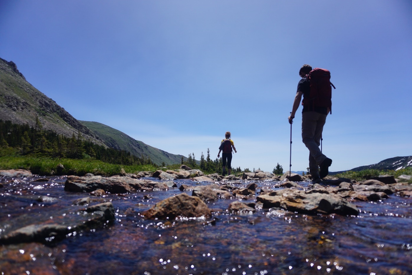 Two hikers crossing a shallow, sunlit stream in a mountainous landscape.