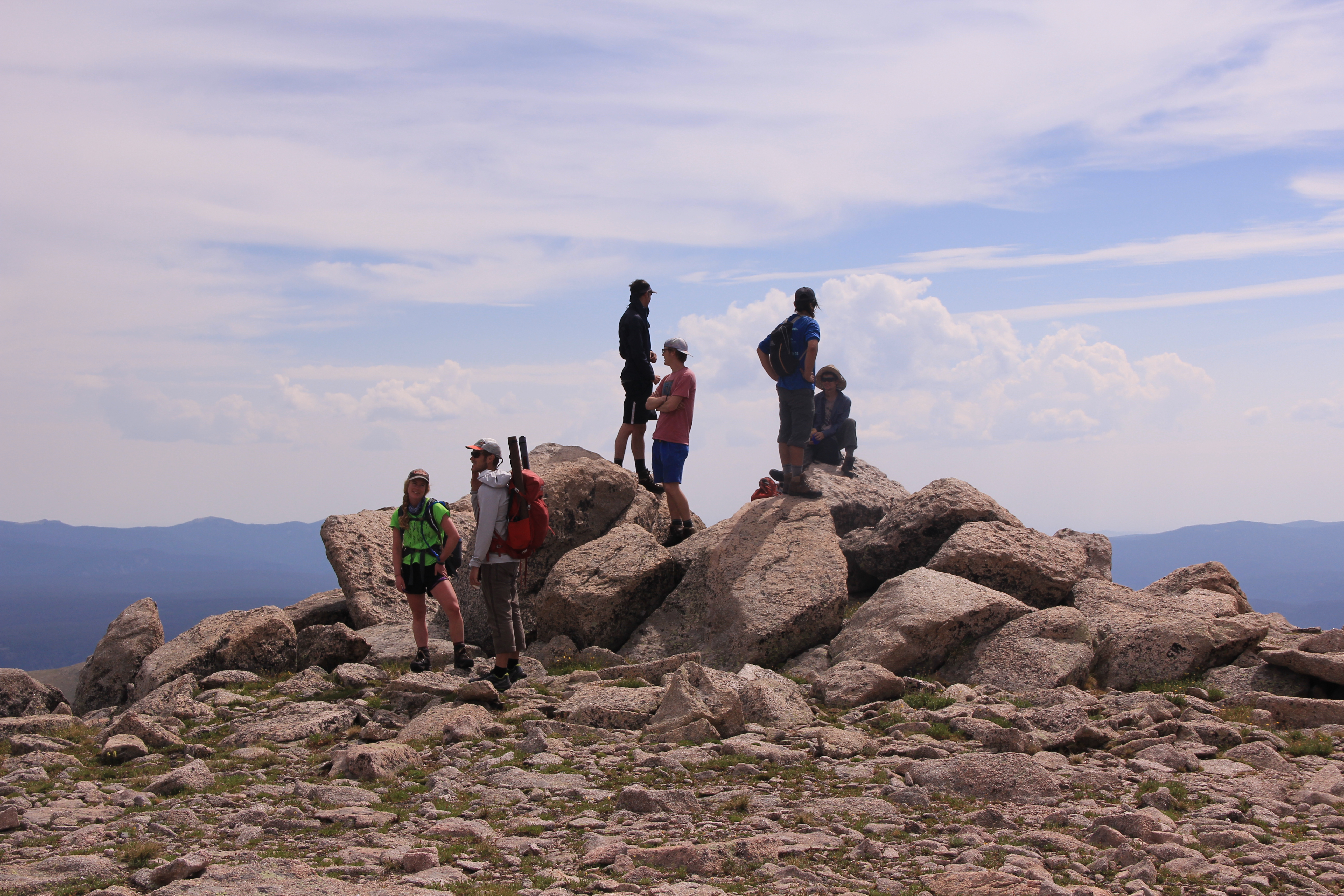 Group of hikers resting on a rocky summit with expansive views of the landscape under a cloudy sky.