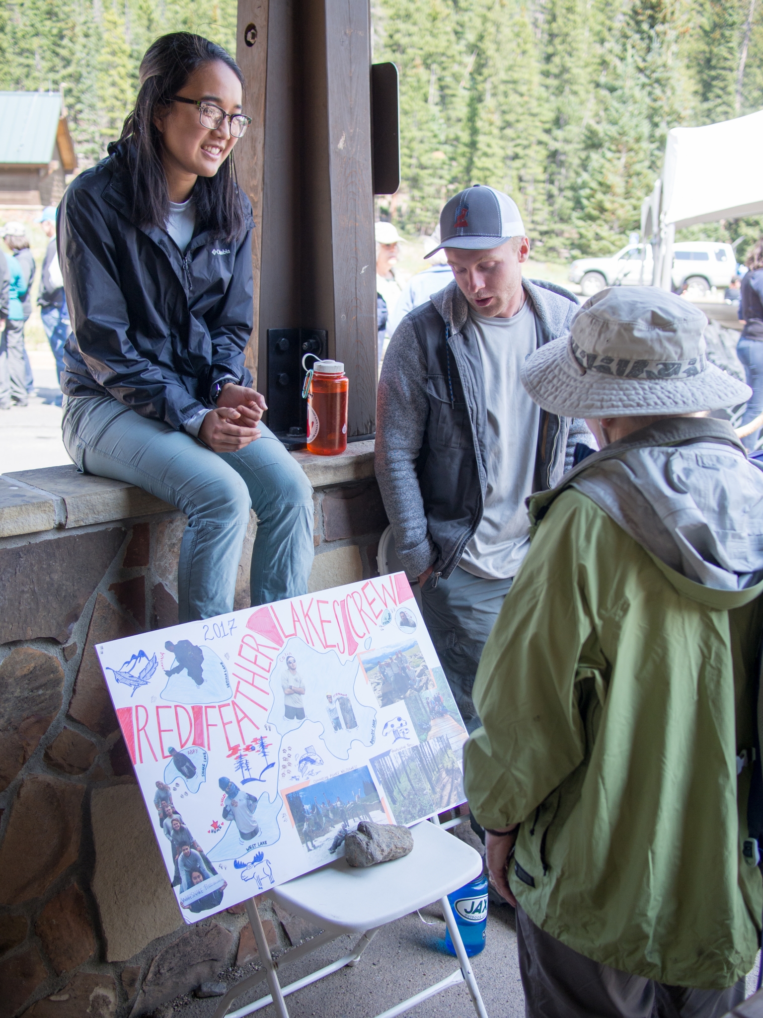 A woman sits on a ledge, smiling and talking to a person wearing a hat, with a sign "redefine lakeshore!" featuring wildlife illustrations, displayed in front of them.