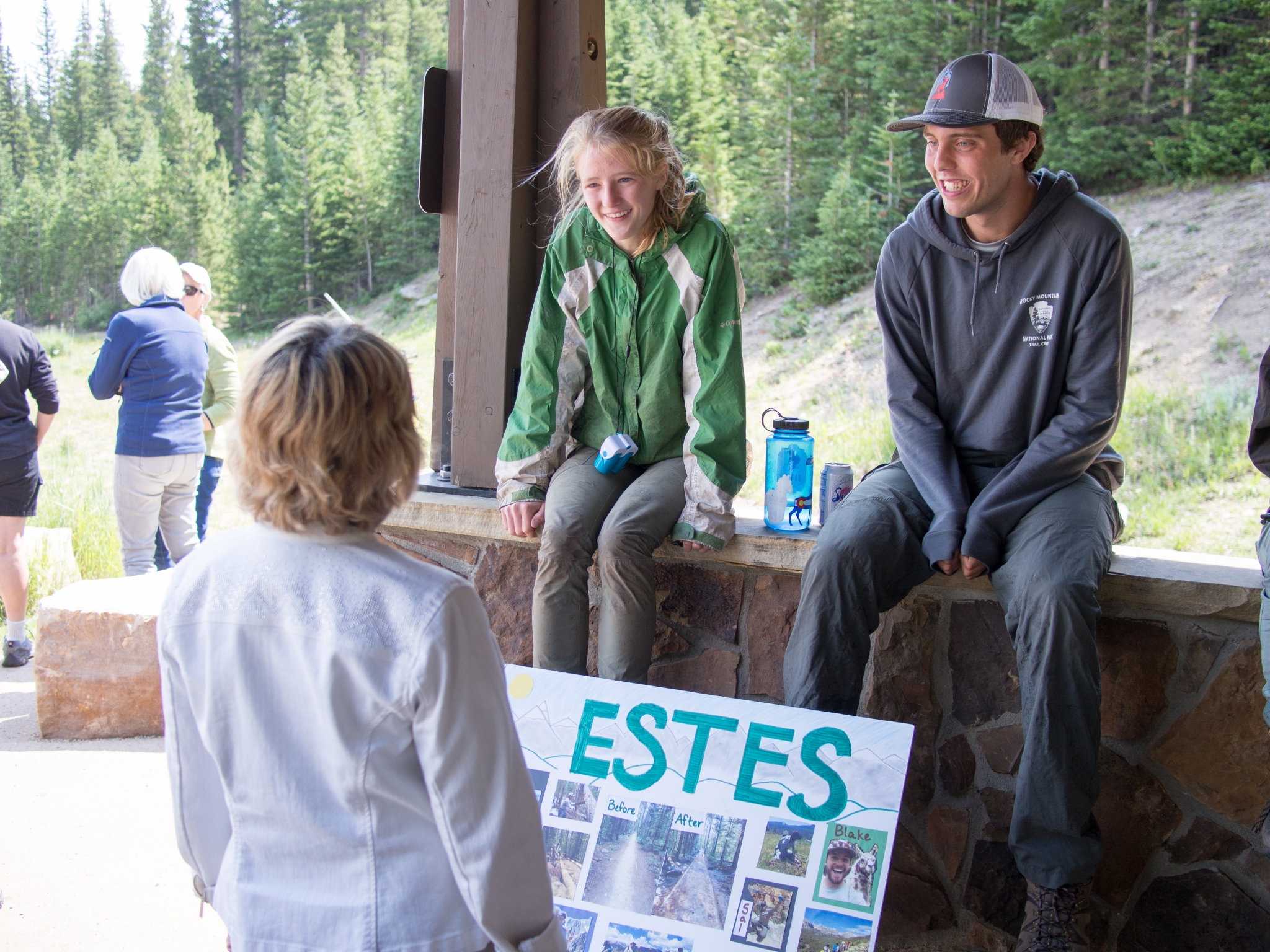 Two park rangers engage with a visitor at an information booth adorned with a sign reading "estes," set against a forest backdrop.