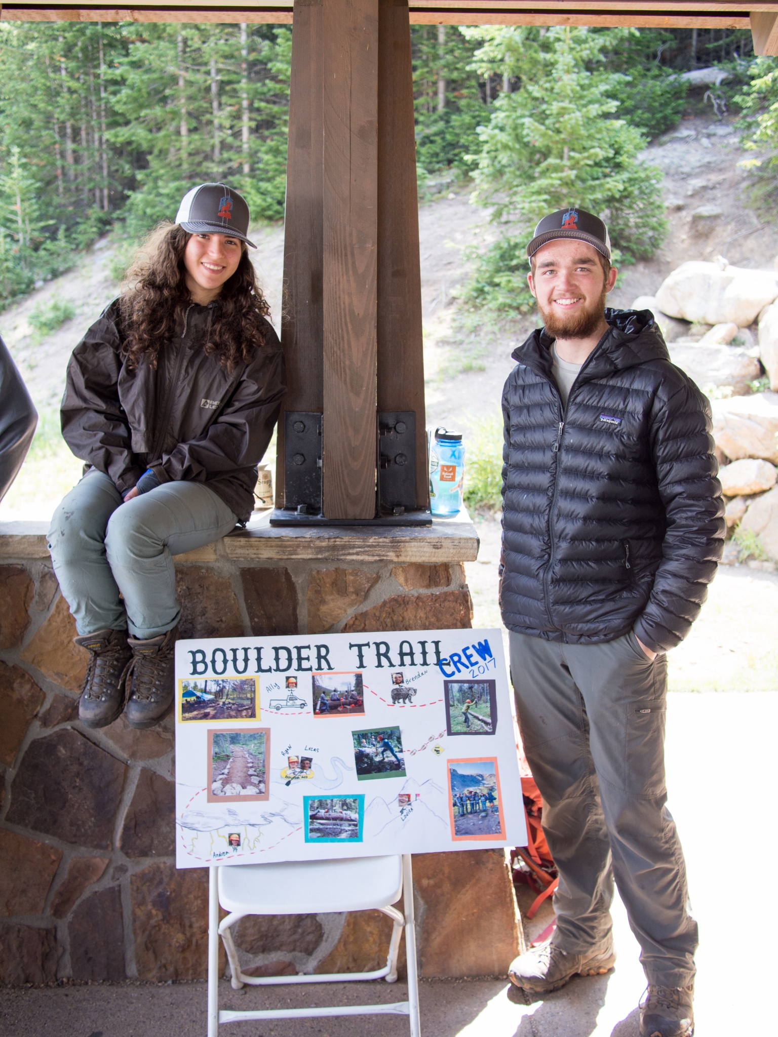 A man and a woman wearing casual outdoor attire and hats, smiling beside a presentation board titled "boulder trail team" at a forested venue.