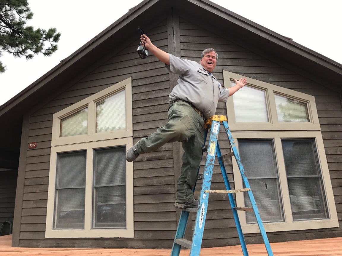 A man on a ladder pretending to fall while holding a hammer, in front of a two-story house with large windows.