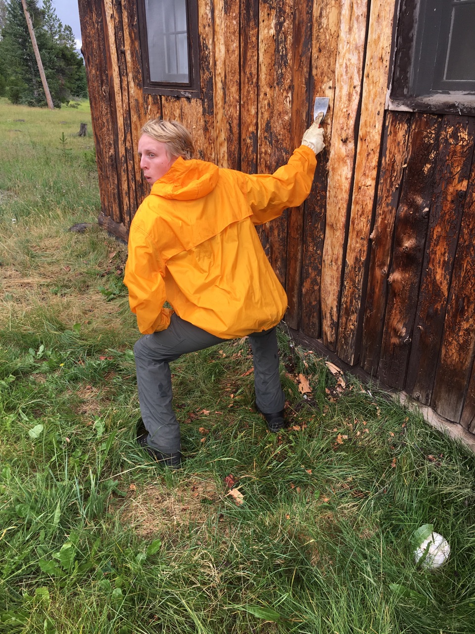 A person in a yellow raincoat pretending to paint a rusty cabin with a brush, green grass and trees in the background.