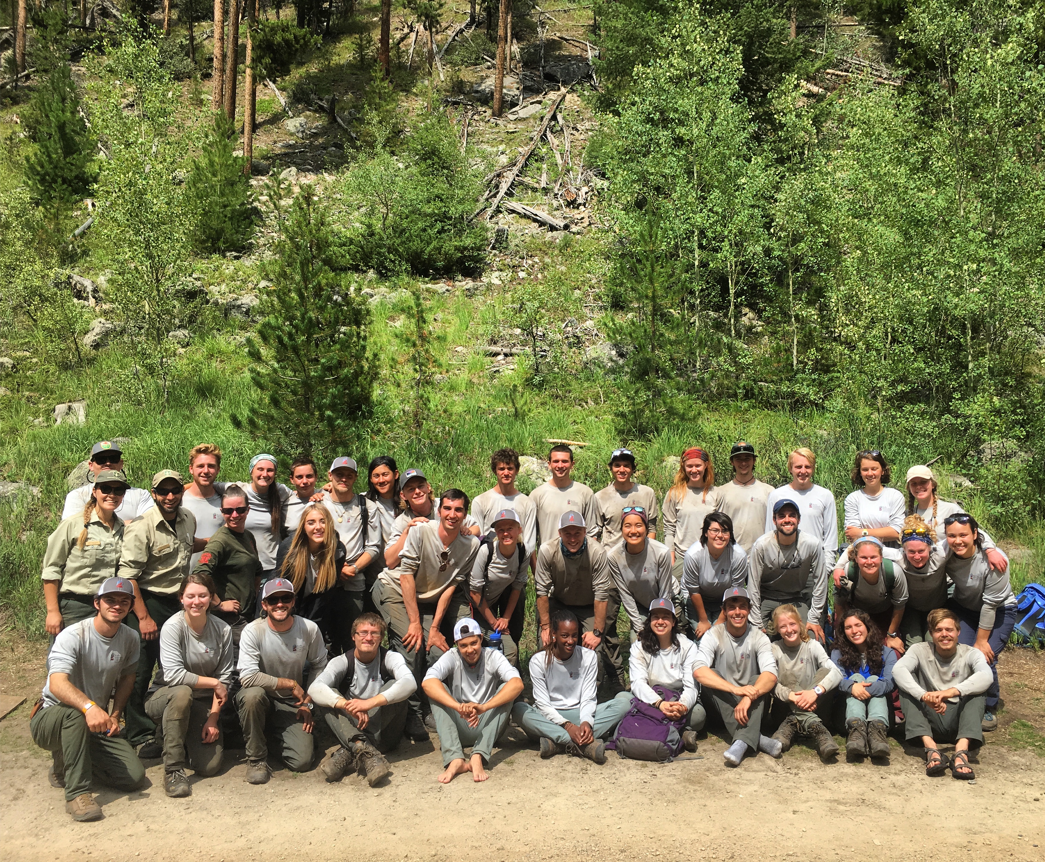 A large group of people in uniforms posing for a photo outdoors, with a forested hillside in the background.