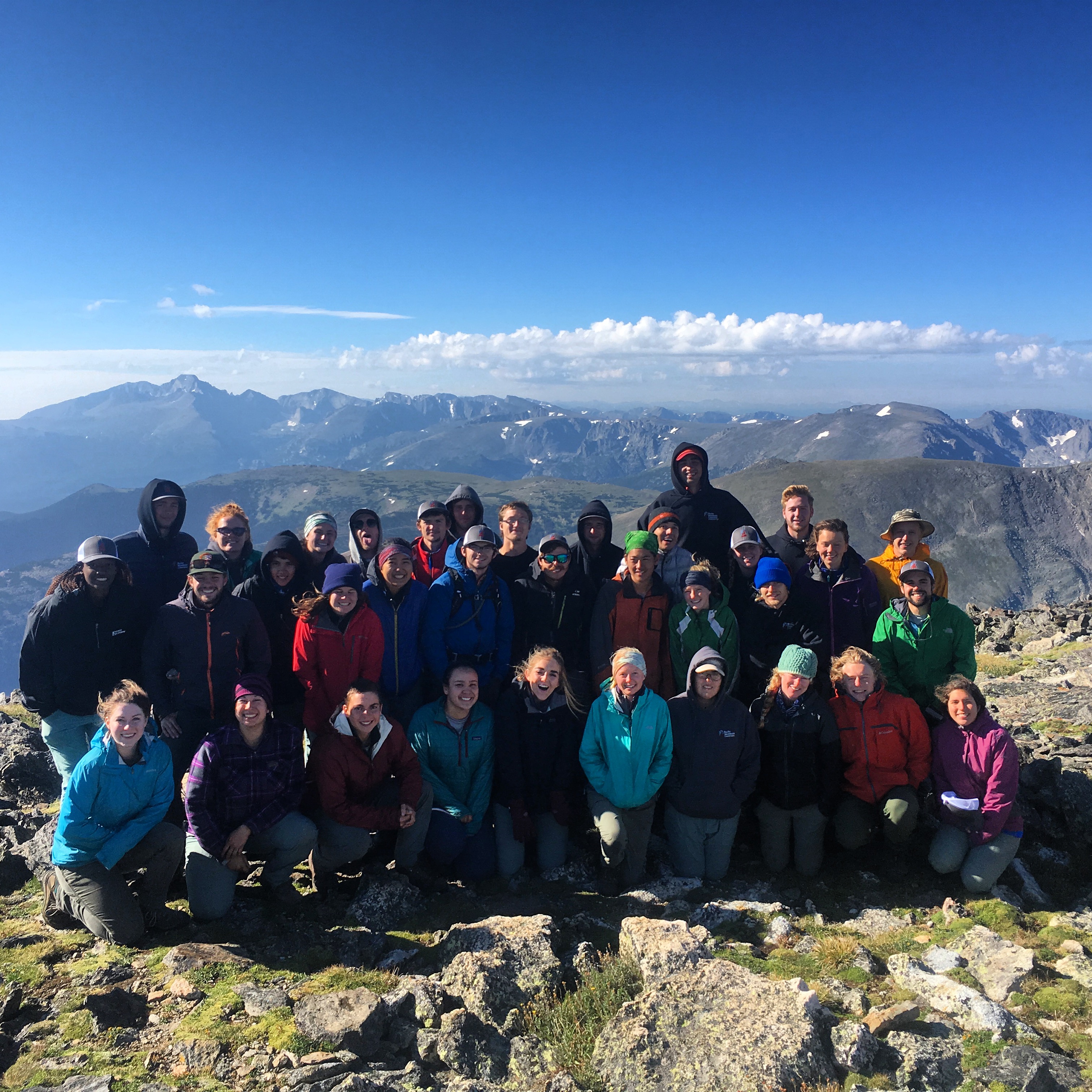 Group of people wearing jackets posing on a mountain summit with clear blue skies and distant mountains in the background.