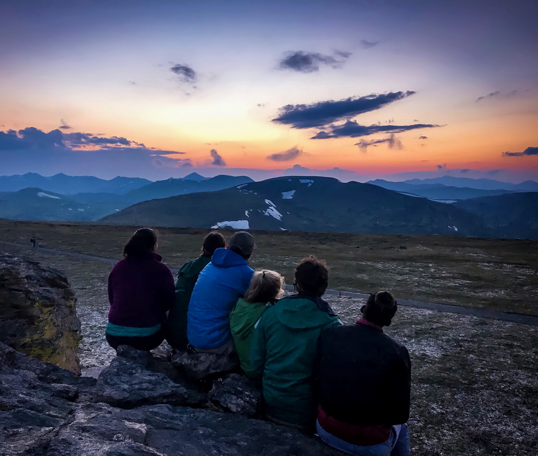 Five people sitting on a rocky edge watching a sunset over a mountainous landscape.