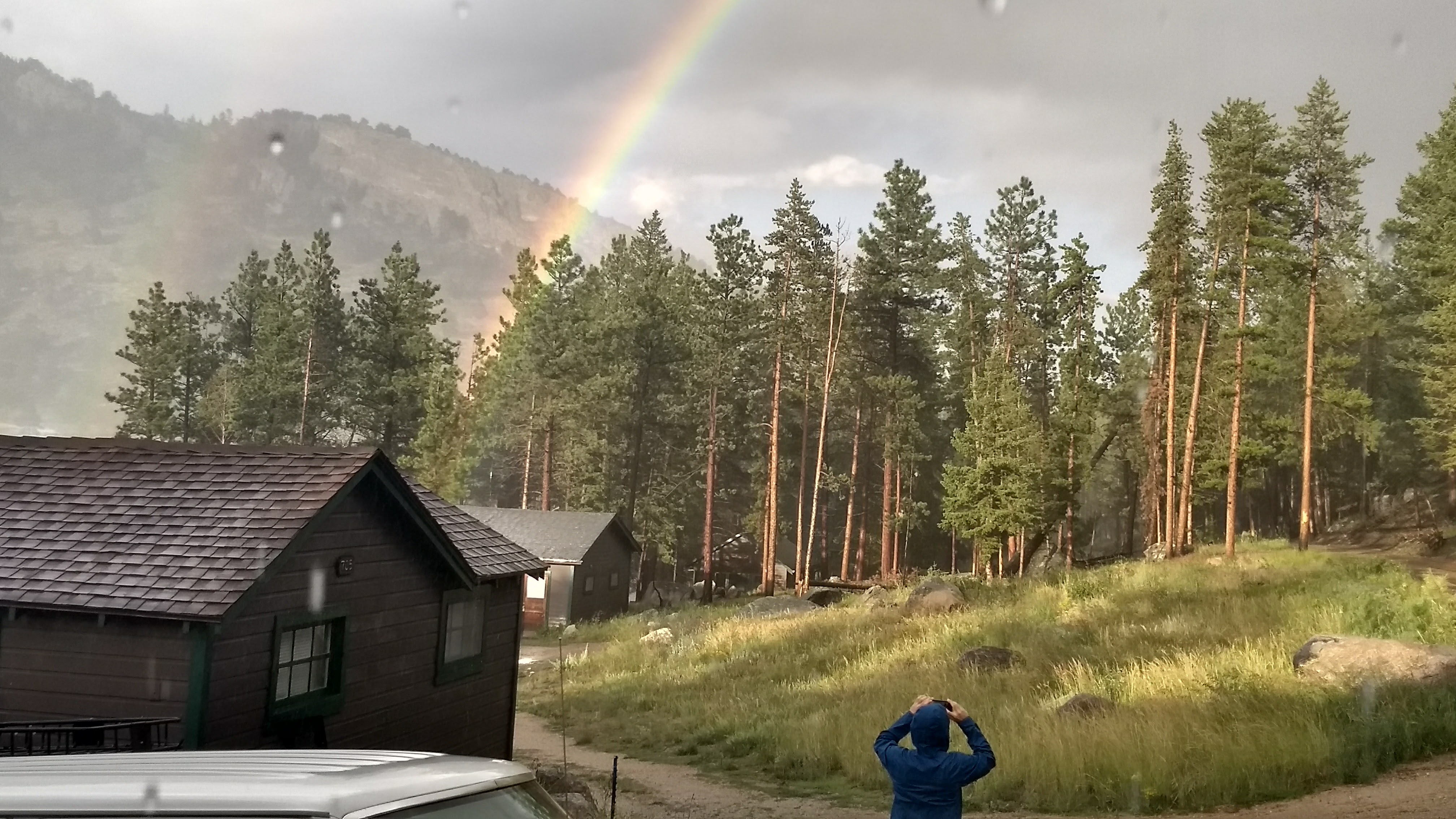 A person photographing a rainbow over a forested landscape with cabins and a cloudy sky.