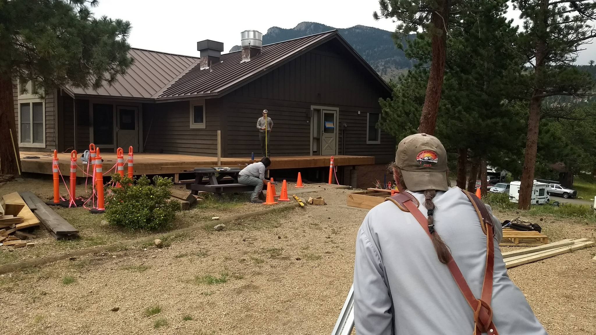 A construction site with workers building a wooden deck in front of a house, surrounded by trees and mountainous backdrop.