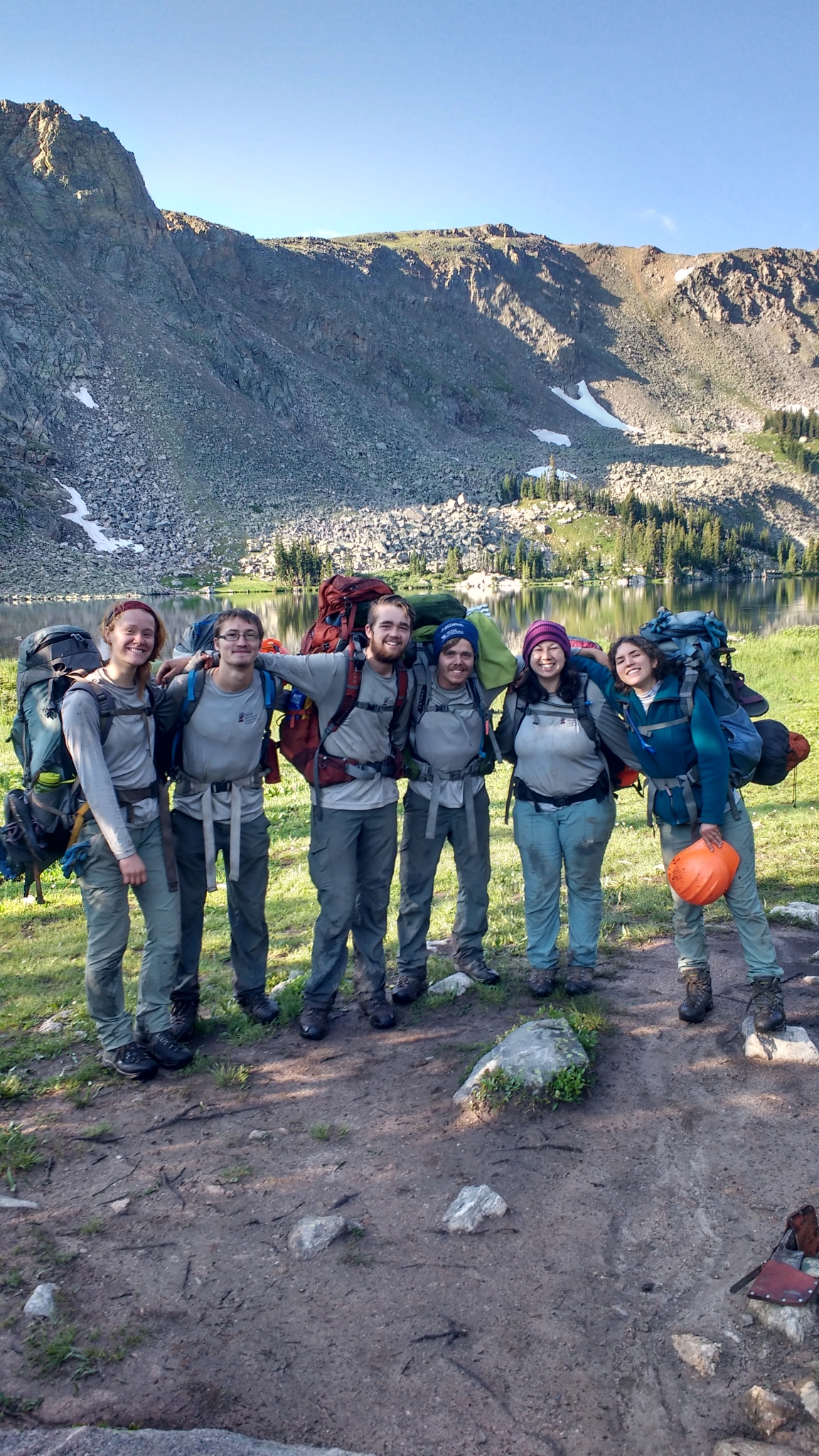 Six hikers with backpacks smiling in front of a mountain lake and rocky terrain under a clear sky.