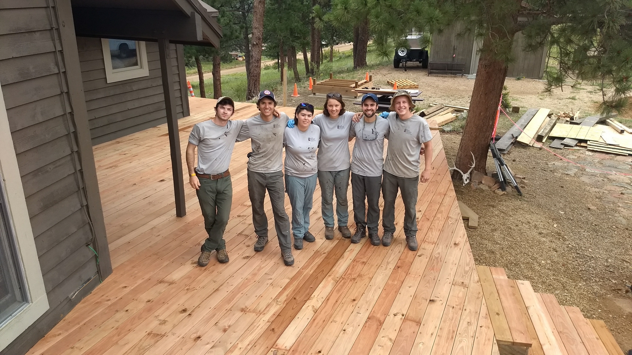 A group of seven construction workers smiling and posing together on a newly built wooden deck, surrounded by building materials and a cabin.